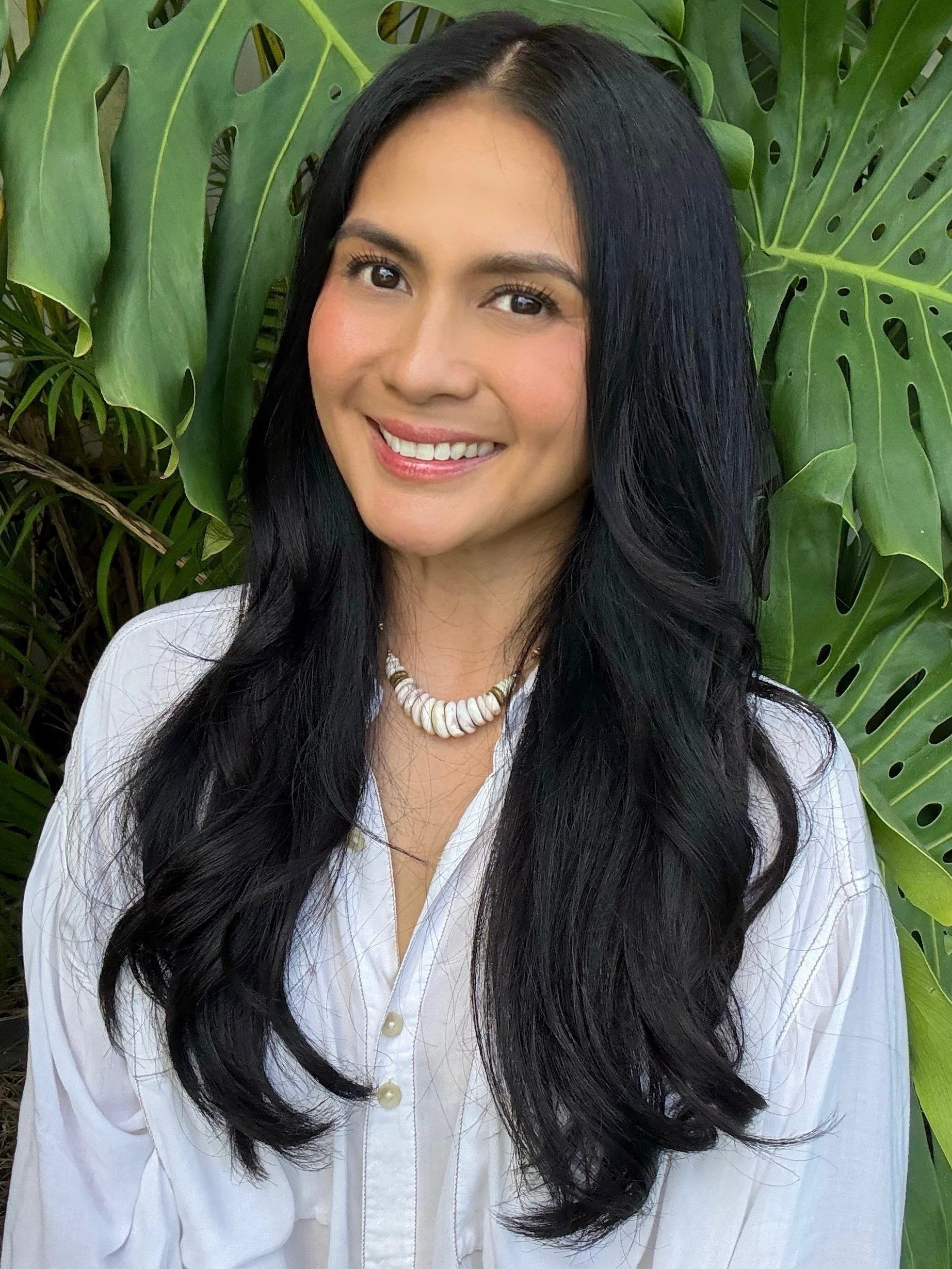 A woman with long black hair, smiling, wearing a white button-up shirt and a shell necklace, standing in front of large green tropical leaves.