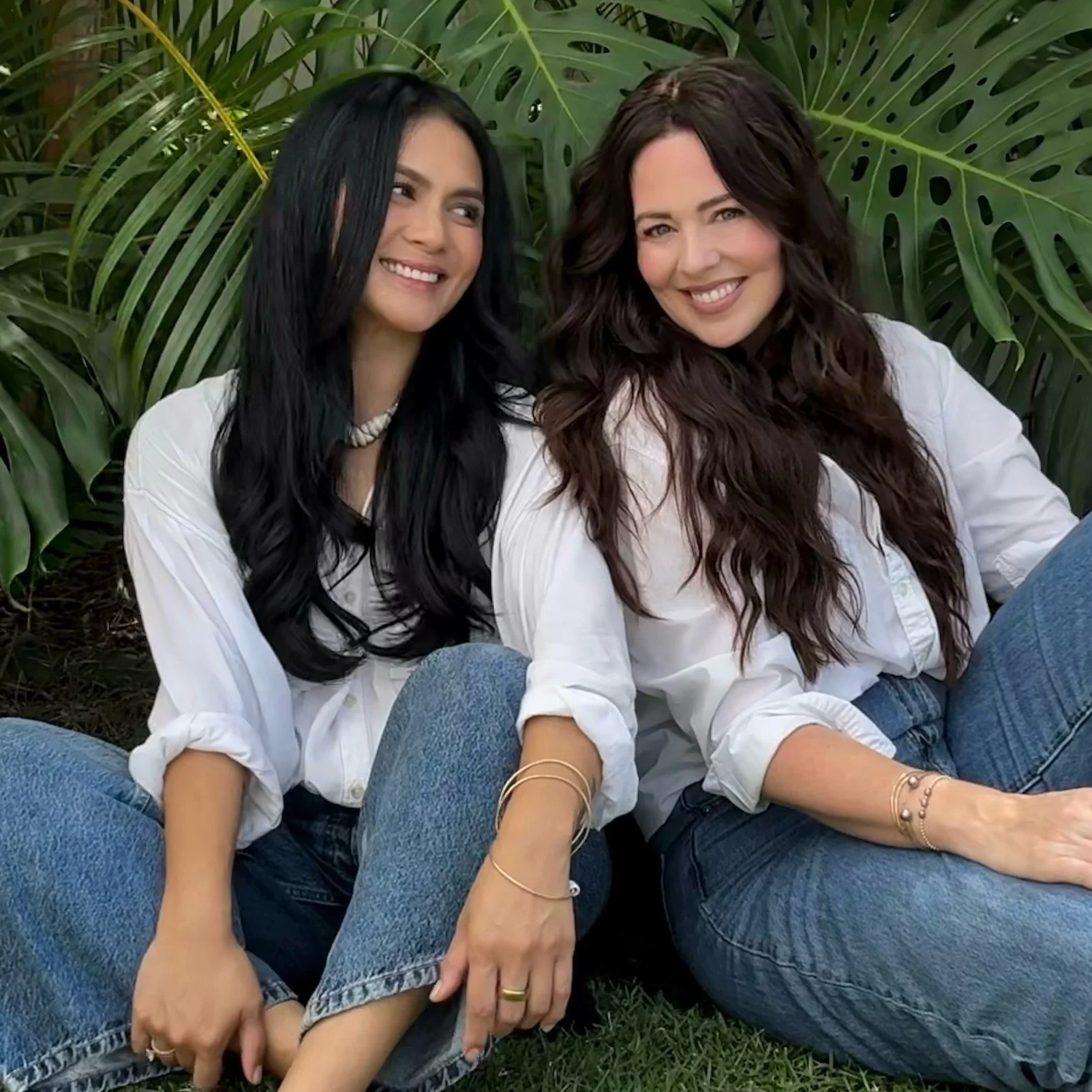 Two women sitting on the grass with large tropical leaves in the background, smiling and wearing white shirts and blue jeans.