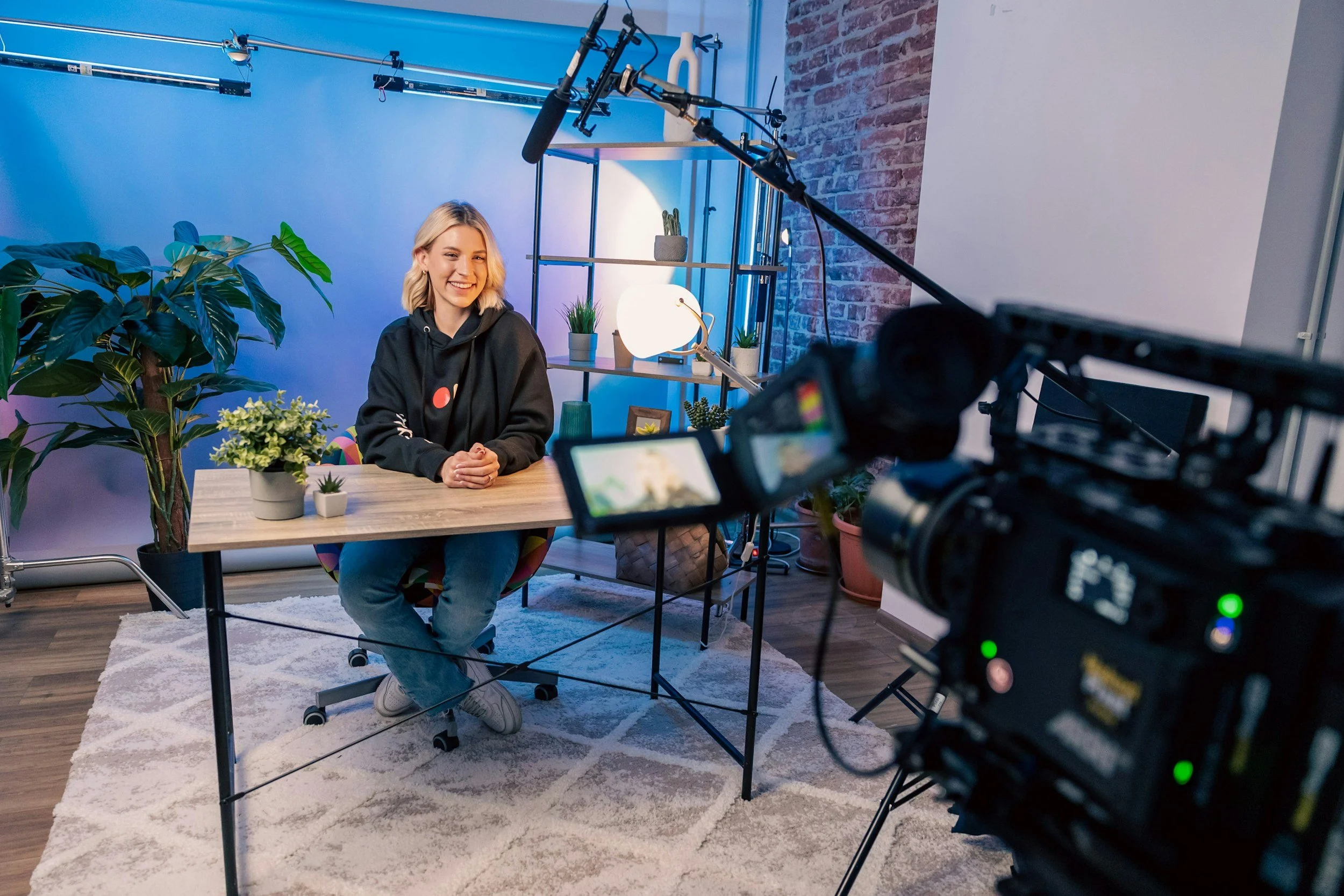 A young woman smiling and seated at a wooden desk during an indoor video recording, surrounded by houseplants, with a brick wall and shelves in the background, as professional camera equipment records her.