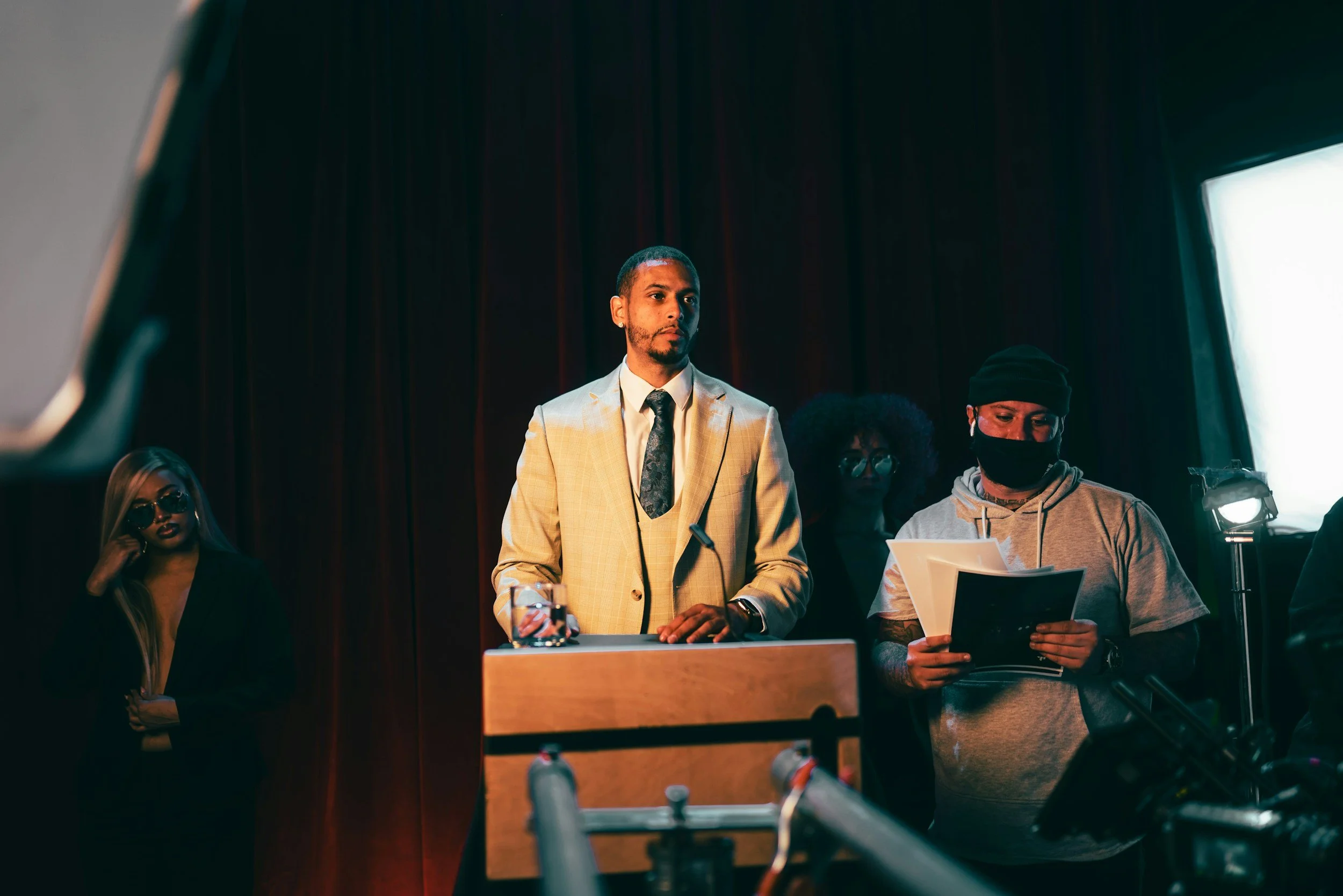 A man in a beige suit and tie stands behind a podium in a dimly lit room with dark curtains. To his right, a person wearing a hoodie and a black face mask holds a paper, and behind them, two women with glasses and dark hair look on. The room appears to be a stage or conference setting.