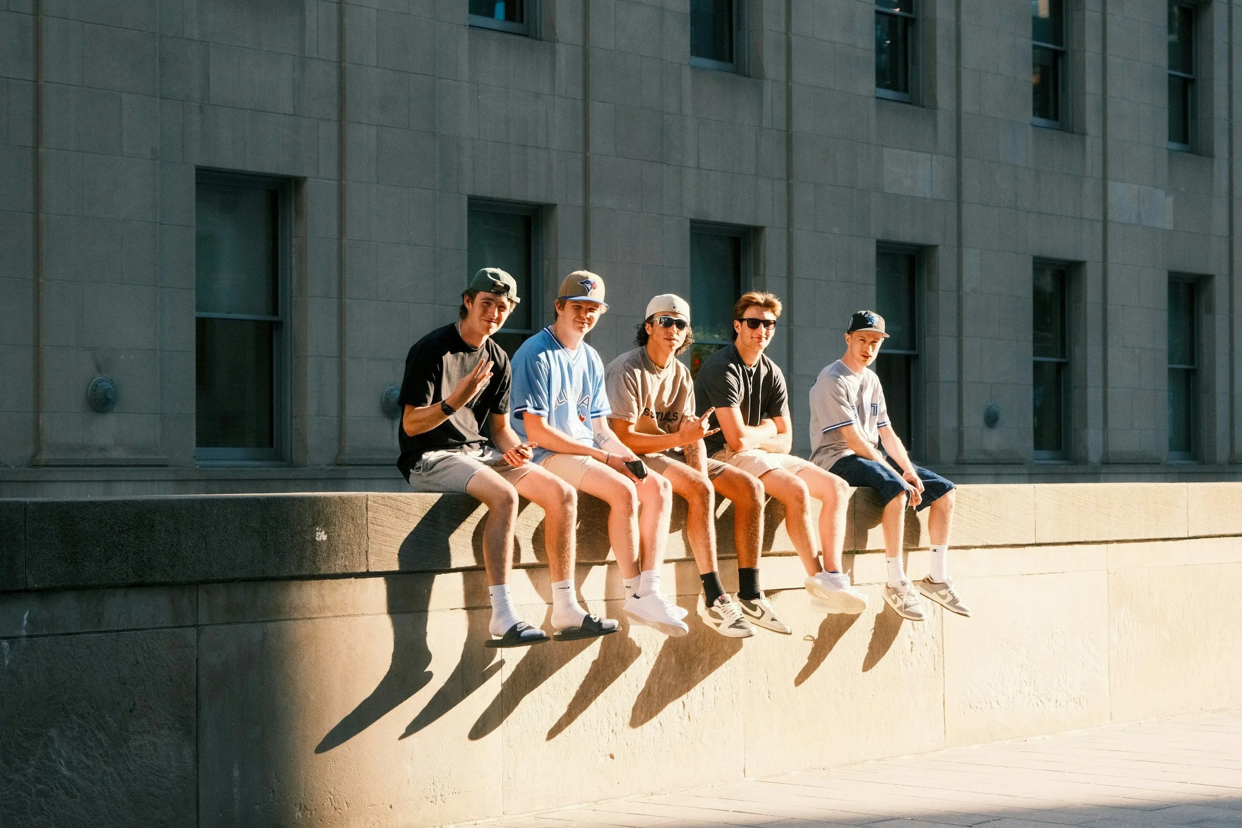 A group of six young men sitting on a ledge in front of a modern building with large windows and stone walls, some wearing baseball caps and sunglasses, in bright sunlight.