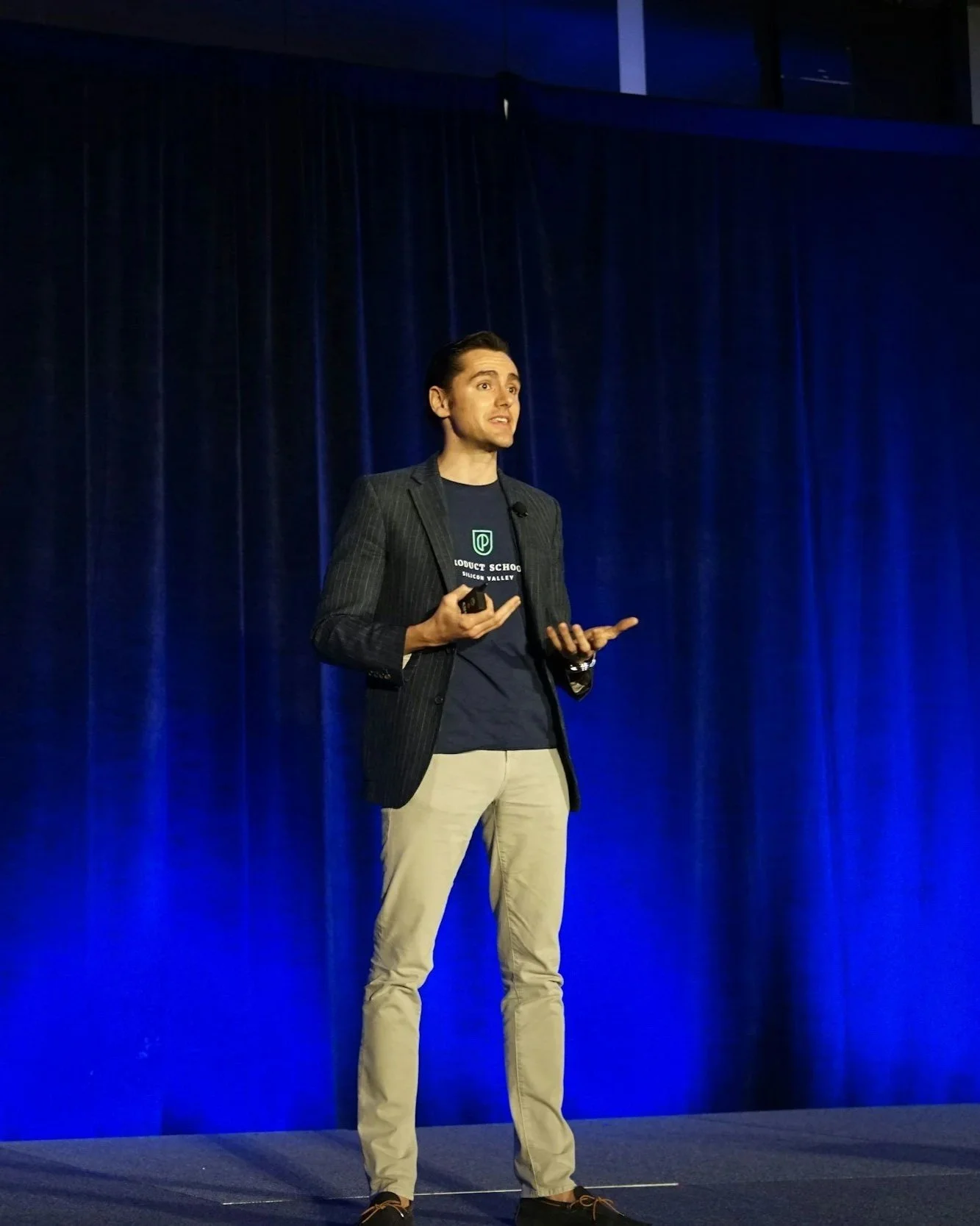 A man standing on a stage with blue curtains, speaking and gesturing with his hands during a presentation or speech.