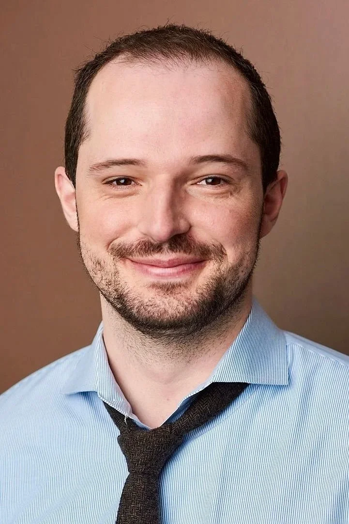 A young man with short brown hair, a beard, wearing a light blue dress shirt and a black tie, smiling at the camera against a brown background.