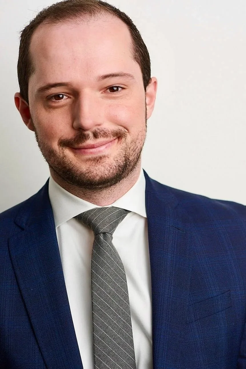 A man in a dark blue suit, white shirt, and gray striped tie smiling in front of a plain white background.