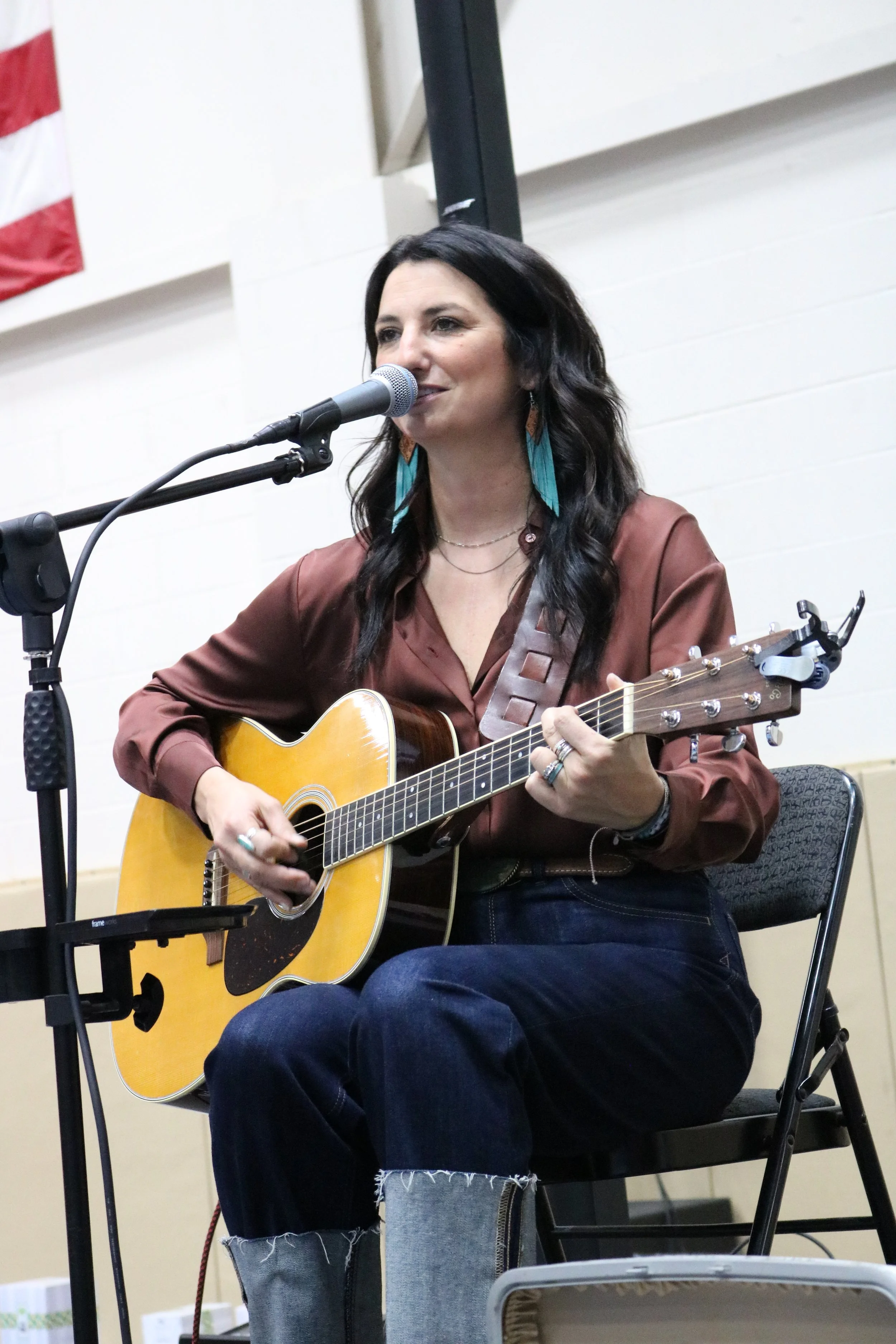 A woman with long dark hair playing an acoustic guitar and singing into a microphone at an indoor event.