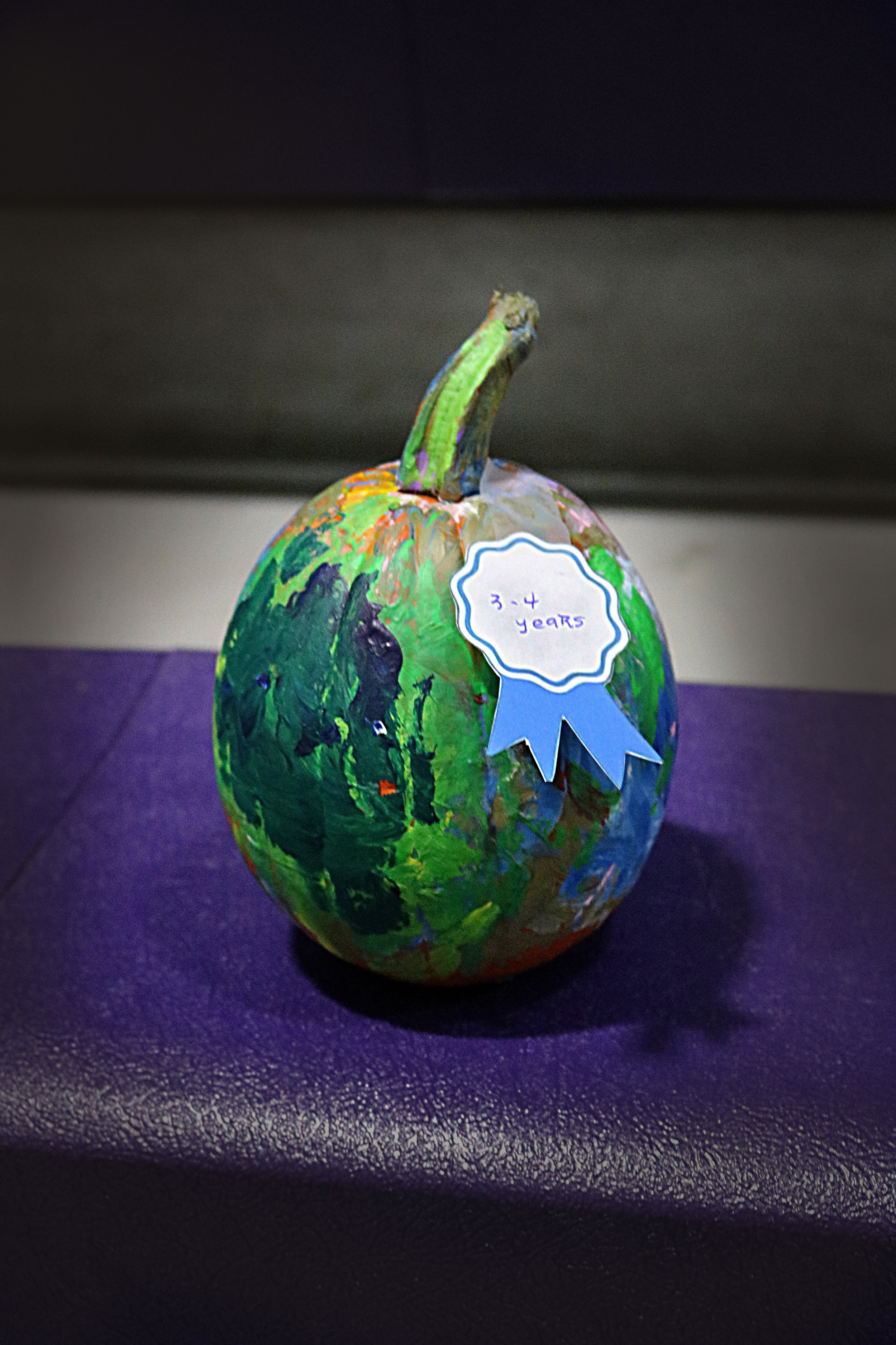 Painted pumpkin with a blue ribbon that reads '3-4 years' placed on a purple surface.