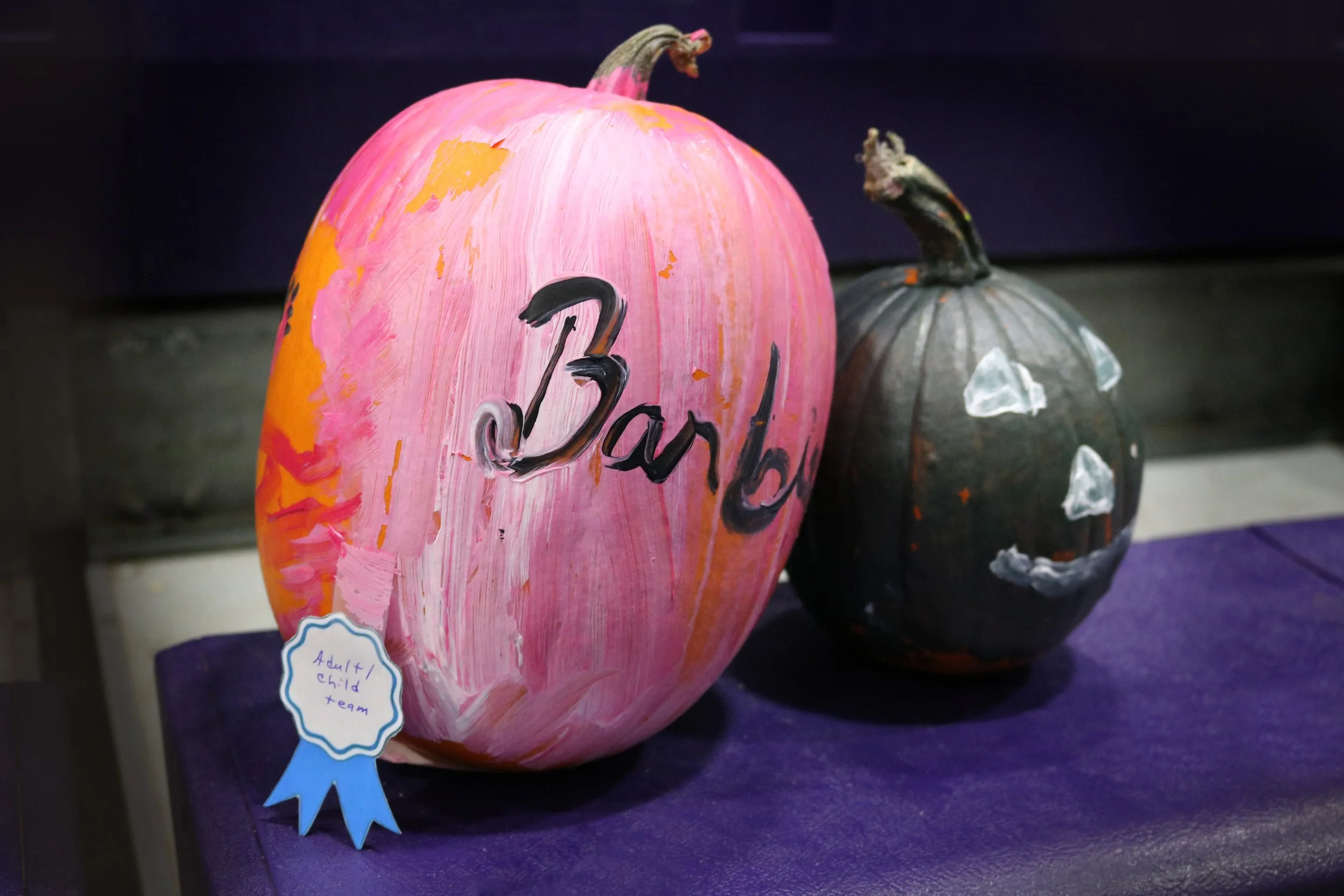 Two decorated pumpkins, one painted pink with the word 'Barb' and a blue ribbon that reads 'Adult/Child Team', and the other painted black with white ghost face details, on a purple surface.