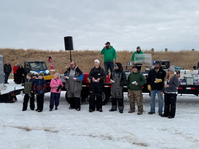 A group of people gathered outdoors on ice, with some holding papers, near a truck with a speaker and equipment, and children dressed warmly, possibly at a winter event or gathering.