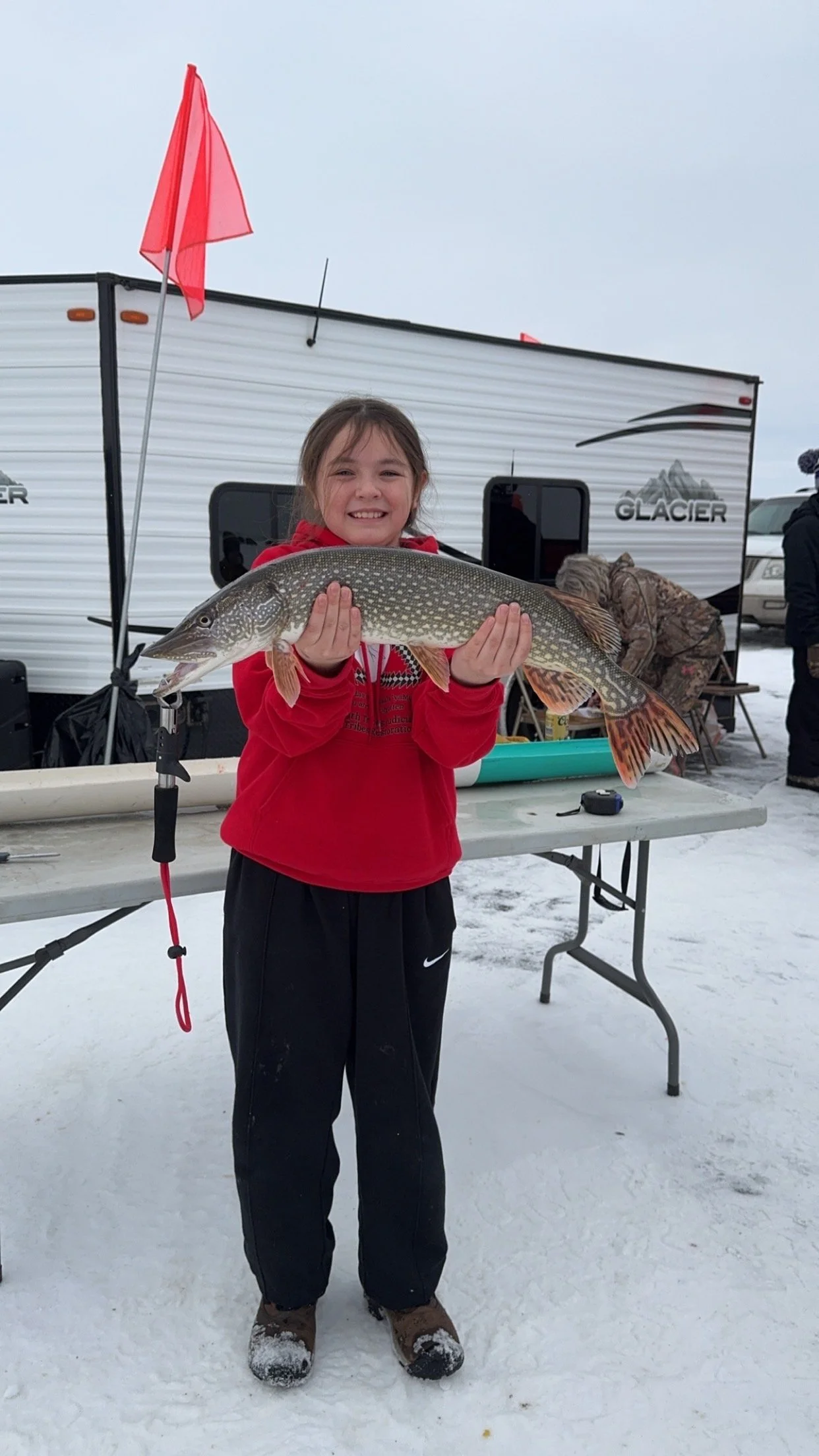 A young girl in a red hoodie standing on snow, holding a large fish she caught, with a camper and other people in the background.