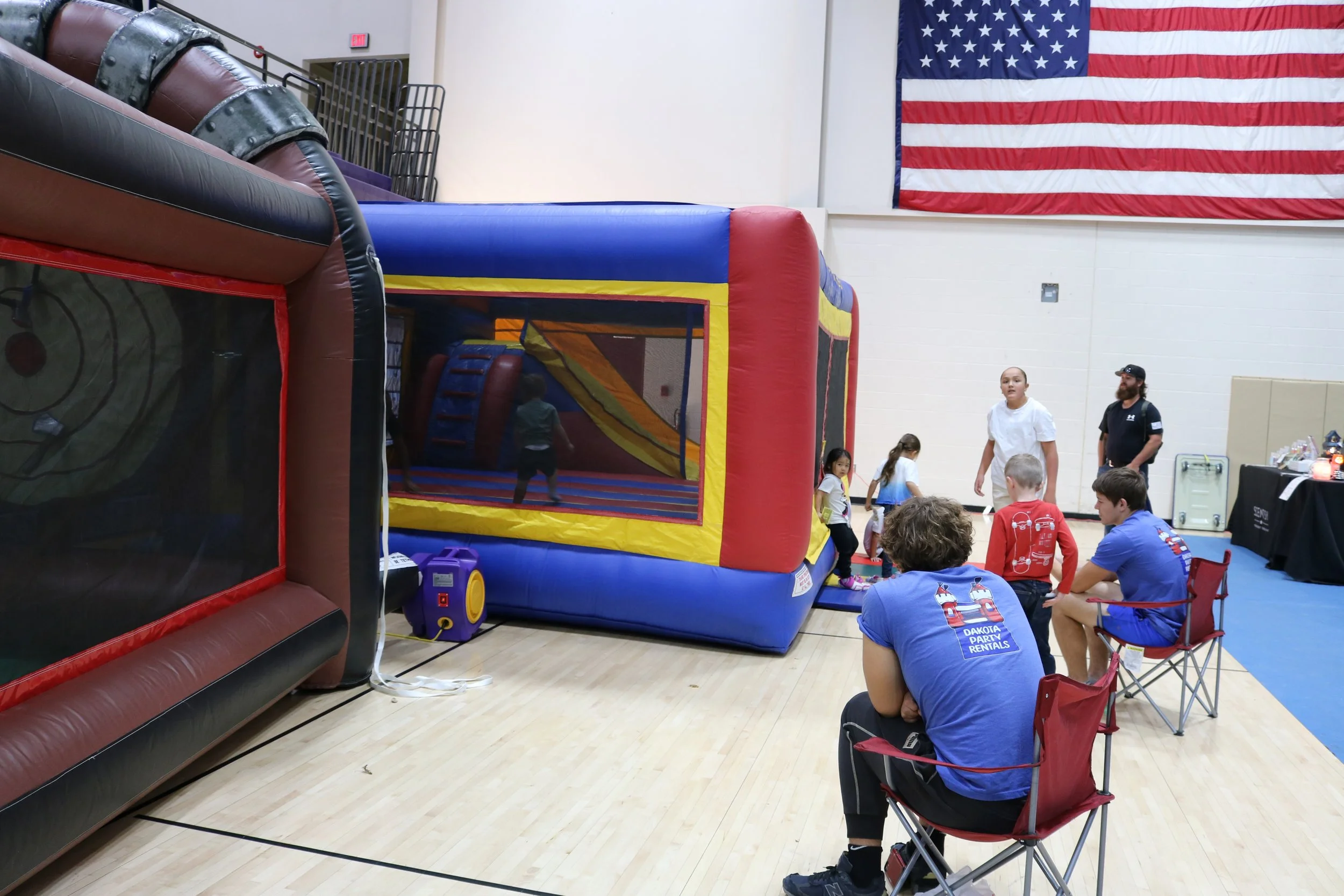 Indoor trampoline park with children playing and adults watching, large American flag hanging on the wall.