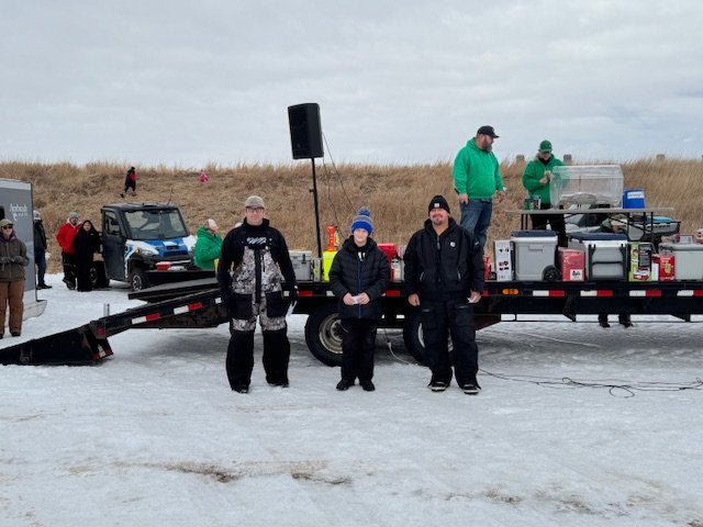 Group of people standing near a flatbed trailer with equipment on a snowy outdoor landscape with grass and cloudy sky.