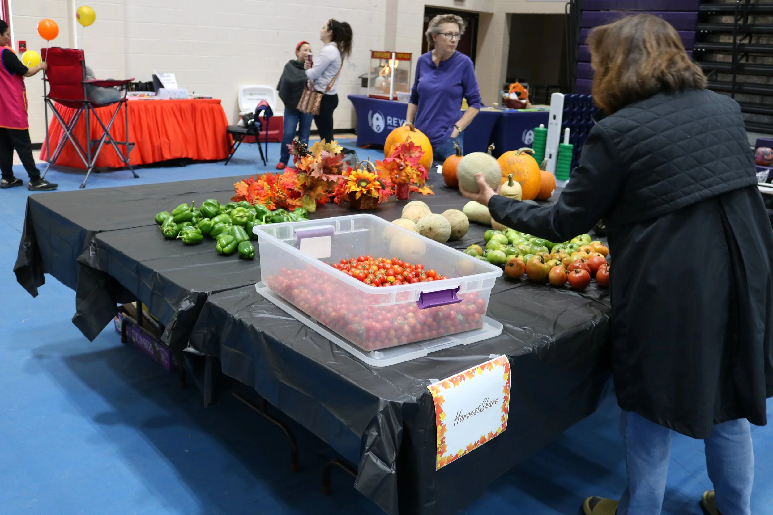 A woman selecting melons at an indoor farmers market table with tomatoes, bell peppers, pumpkins, and fall decorations, and other people shopping in the background.
