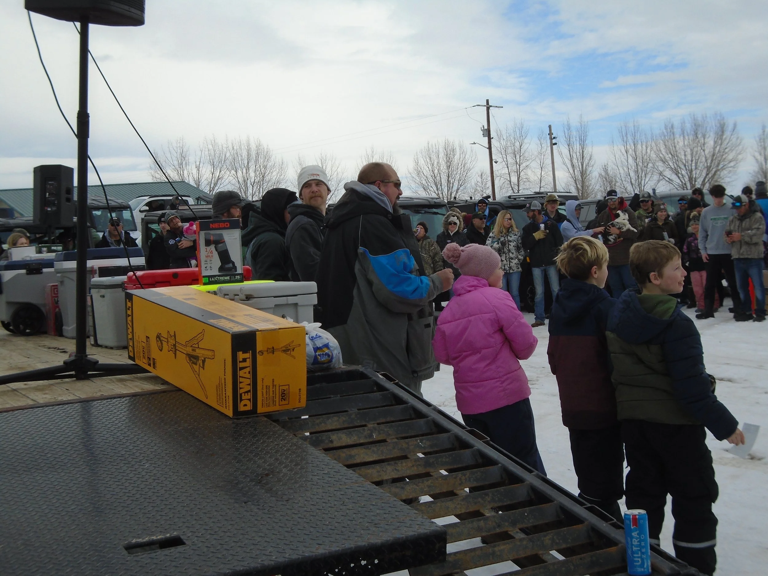 A crowd of people gathered outdoors on snow-covered ground, some are watching and others are talking. There are tables loaded with equipment and supplies, including a DeWalt tool box and a NEBO flashlight. Some children and adults are wearing winter clothing, and the sky is partly cloudy.