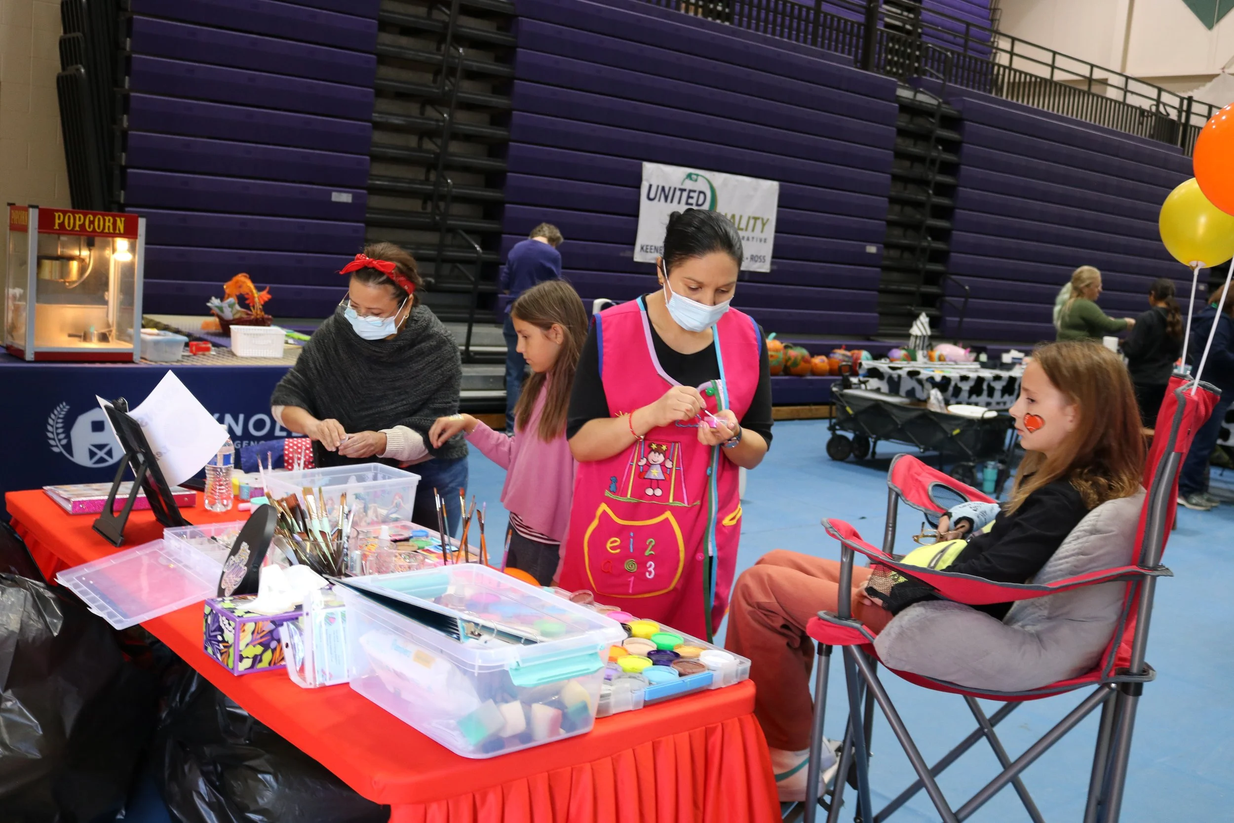 A girl with face paint sitting in a red and gray camping chair, at a face painting booth at a community event. Two women are working at the booth, one in a pink apron and the other in a gray shawl, both wearing masks. The table is filled with face paint supplies.