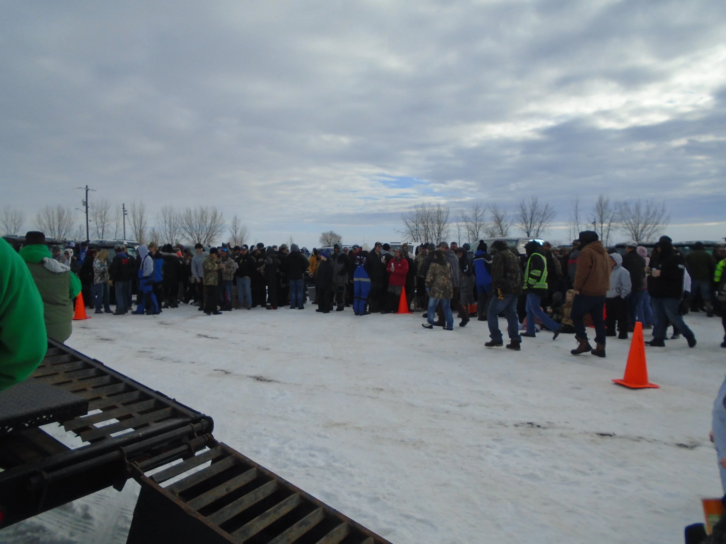 Crowd of people standing in a line on a snowy day, with a few orange cones marking the area, and a cloudy sky overhead.