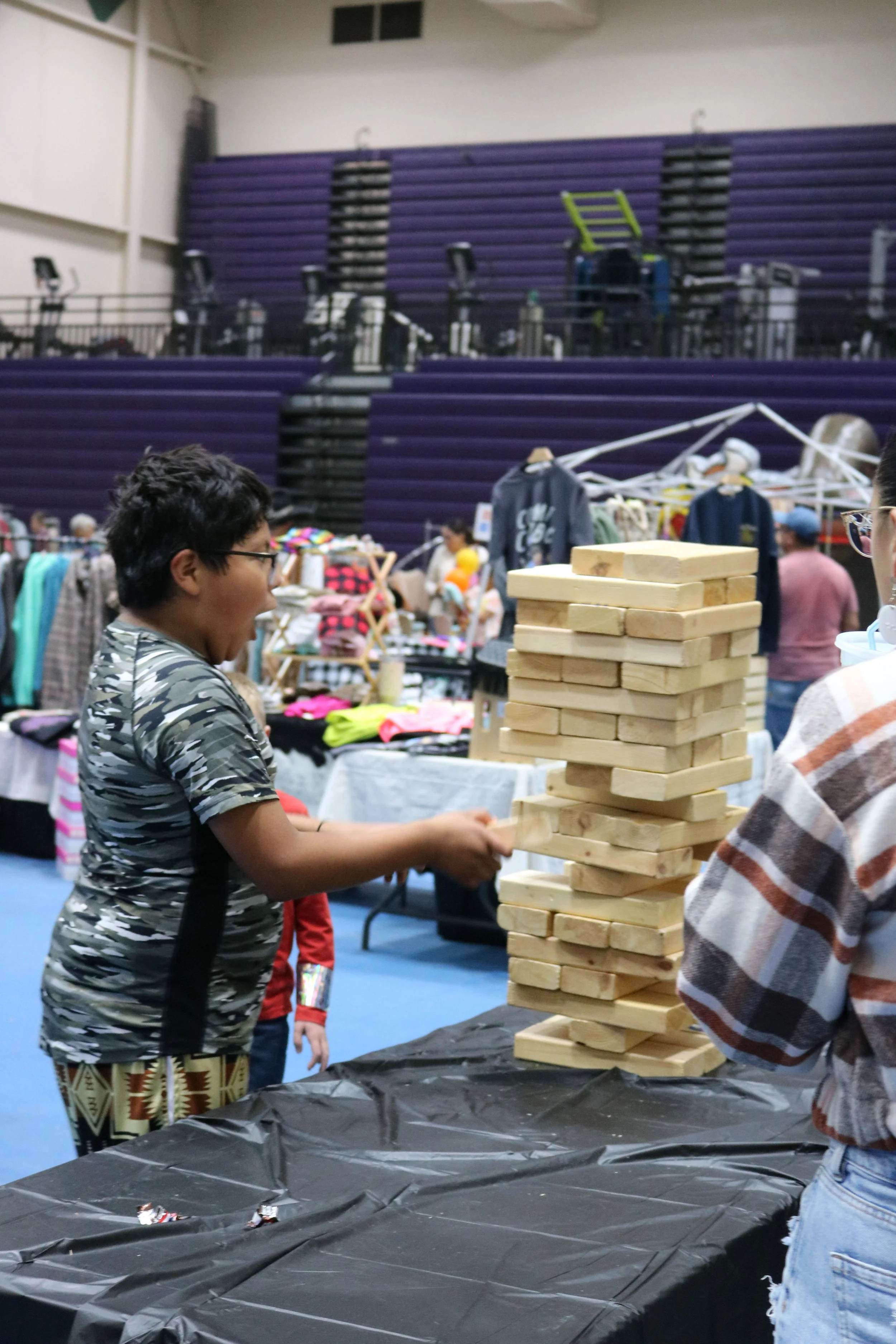 A boy playing Jenga at an indoor market or fair, with tables of clothing and other stalls in the background.