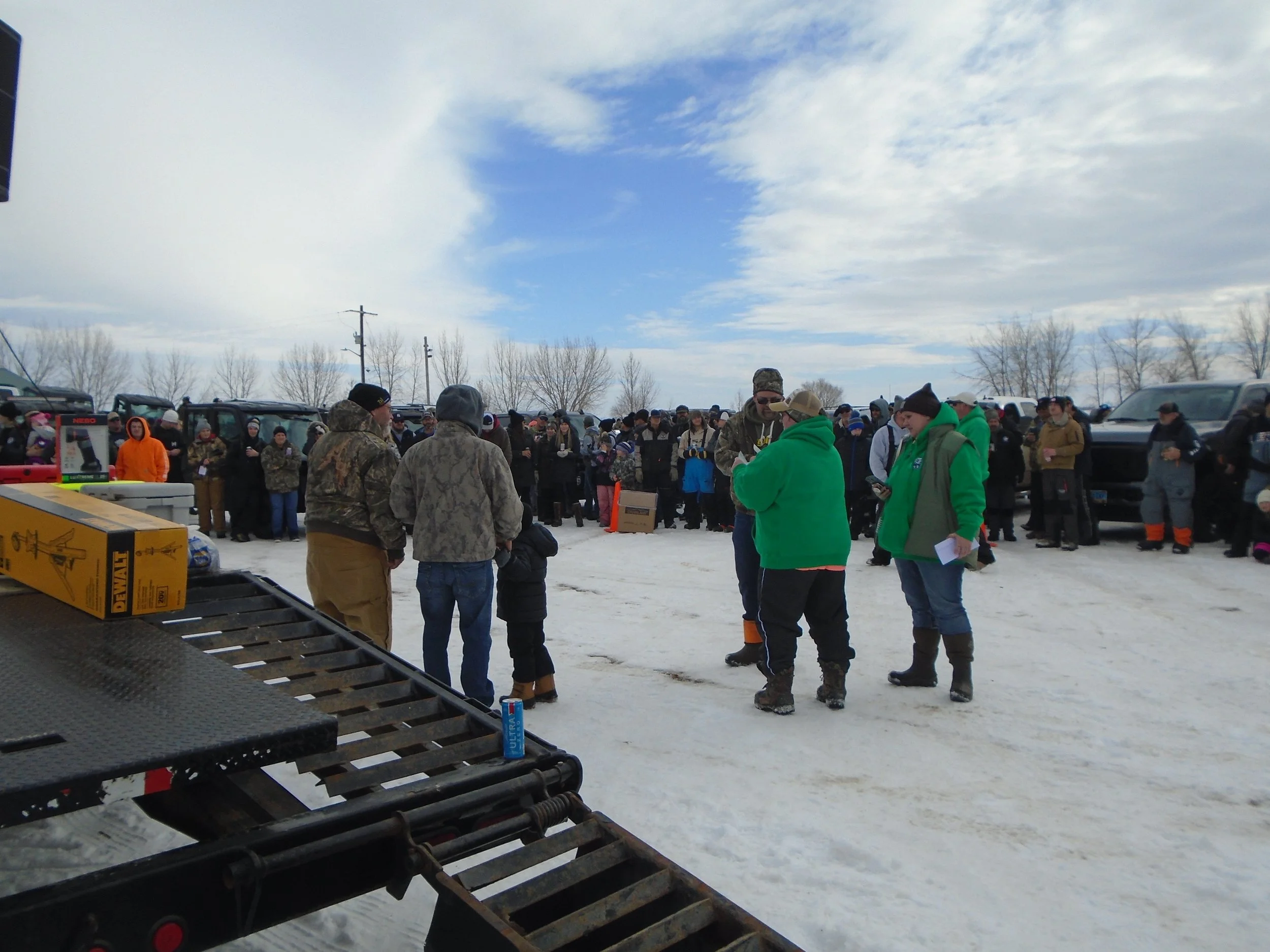 People gathered outdoors on snow-covered ground, some wearing warm clothing, in a line or circle, with vehicles parked in the background and a cloudy sky overhead.