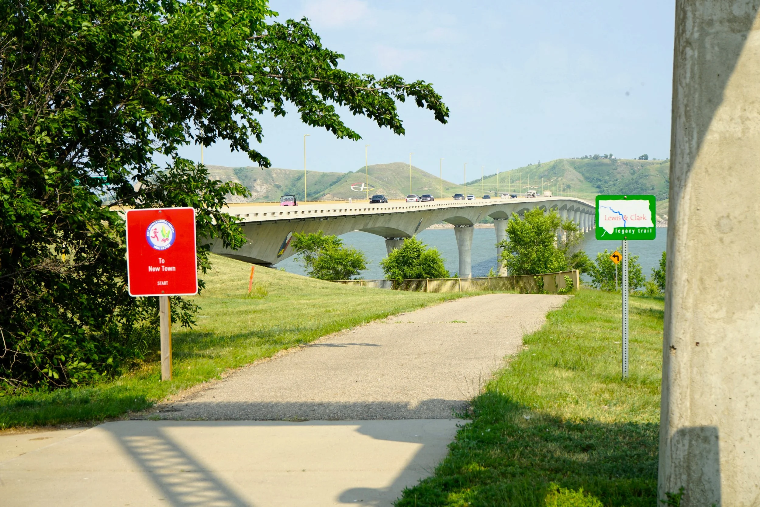 A paved pathway with green grass on both sides leads towards a bridge over a body of water, with hills in the background. There are two signs along the path, one red and one green, indicating directions and trail information.