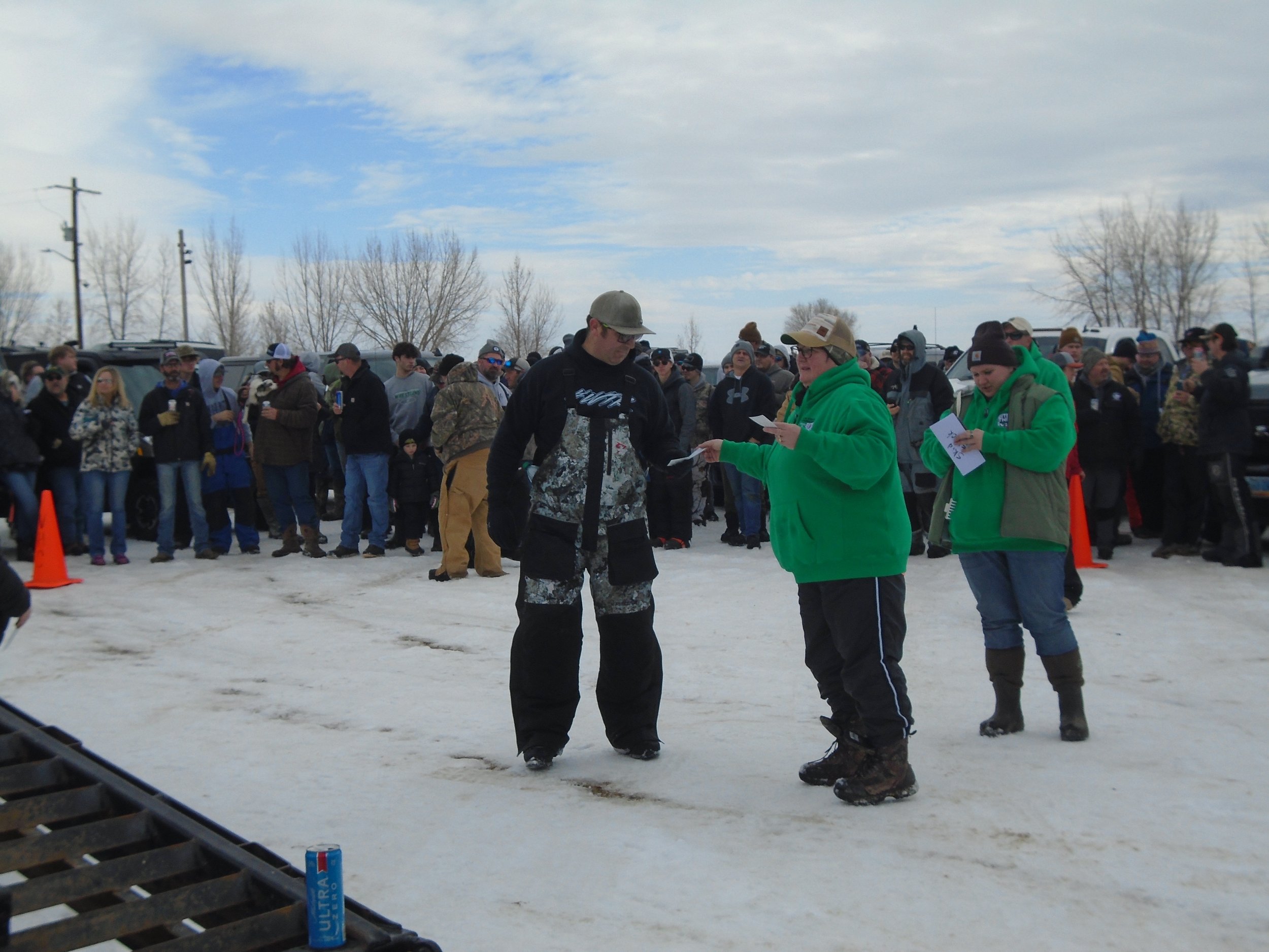 People gathered outdoors on snowy ground, some wearing winter clothing, participating in or observing an event, with a man and two women in the foreground reading papers.