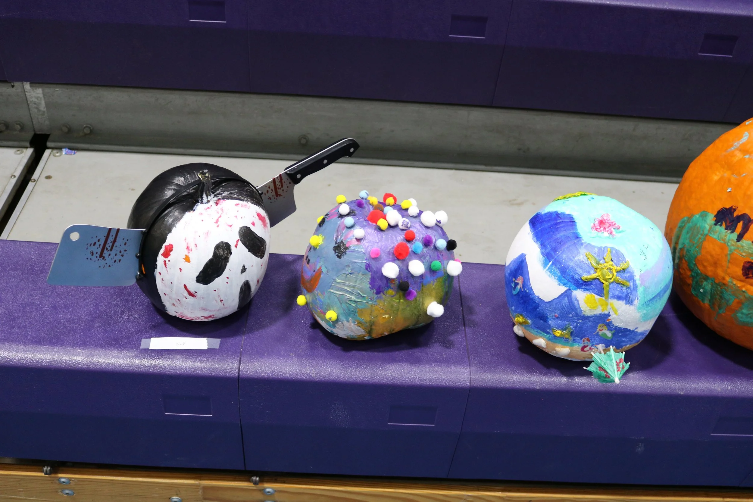 Three painted pumpkins displayed on a purple surface, decorated with various colors and materials, with a knife and painter's tools nearby.