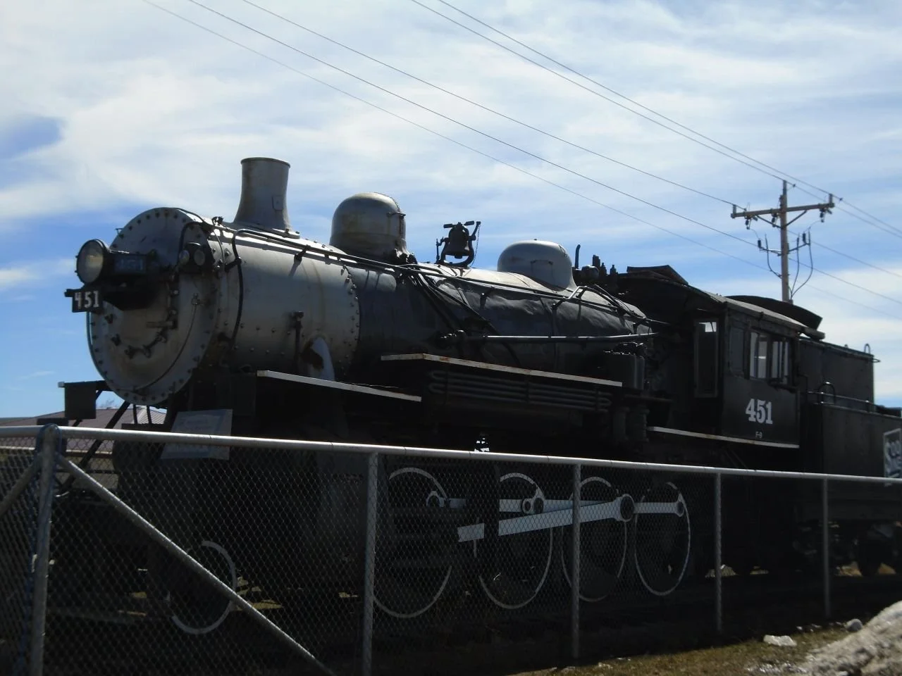 An old black steam locomotive, numbered 451, displayed outdoors behind a chain-link fence, with telephone poles and a partly cloudy sky in the background.