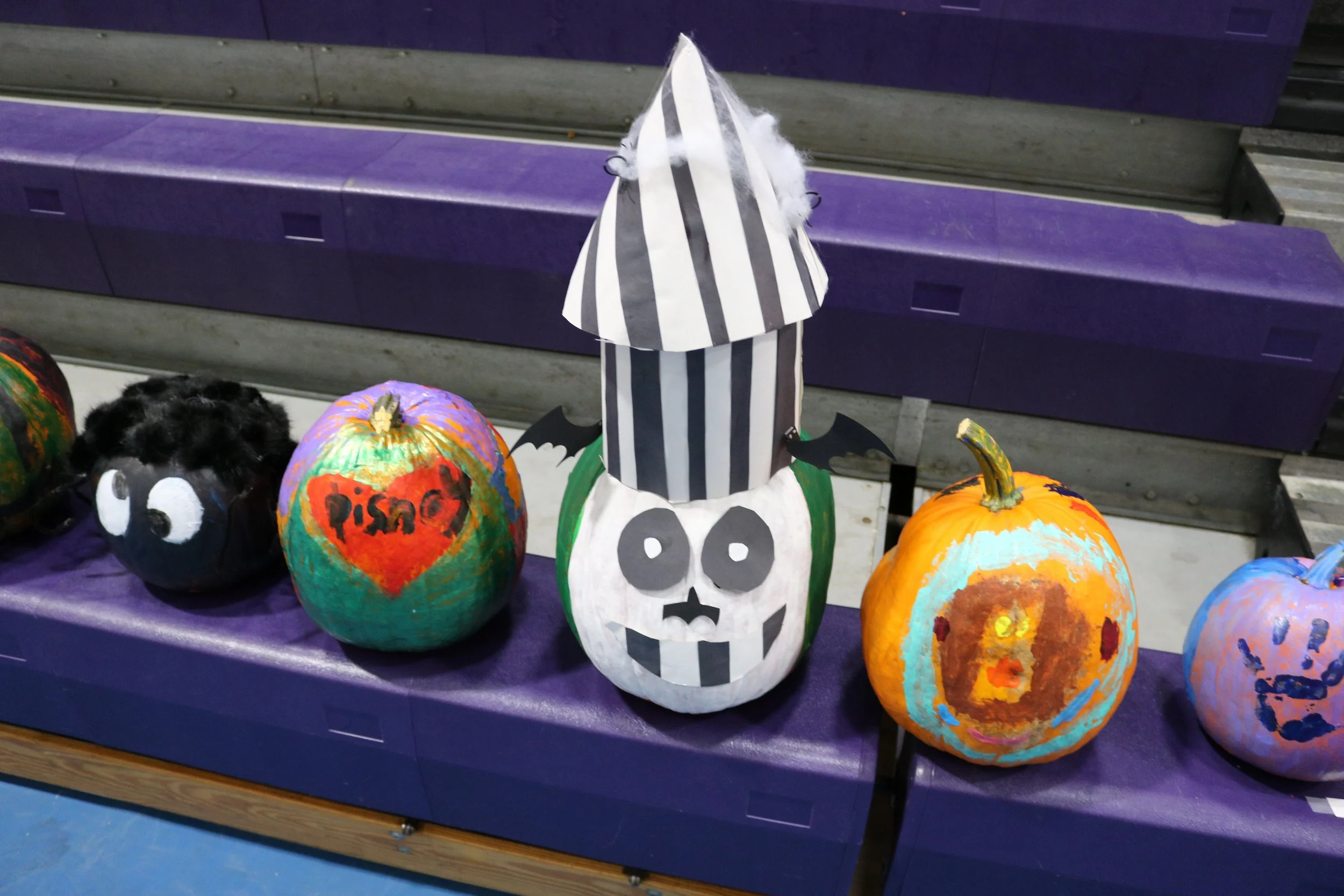 Decorative pumpkins on a purple display table, painted with Halloween themes, including one with a skull, one with a pumpkin face, and one with a black face with eyes. A Halloween-themed paper mache decoration with a skull face and a striped witch hat is in the center.