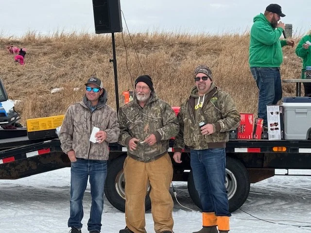 Three men in winter clothing standing on snow next to a black trailer with outdoor equipment, celebrating outdoors with drinks, while two more people are in the background near a table.