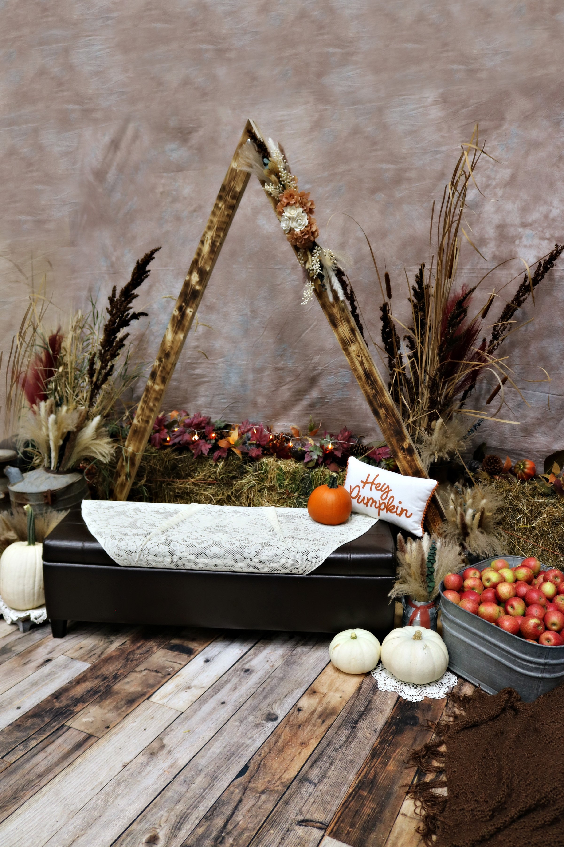 Autumn-themed indoor display with a black bench, a lace cloth, small pumpkins, apples, a pillow saying 'Hey Dumpling,' dried grasses, a decorative triangle frame, and rustic decorations on a wooden floor.