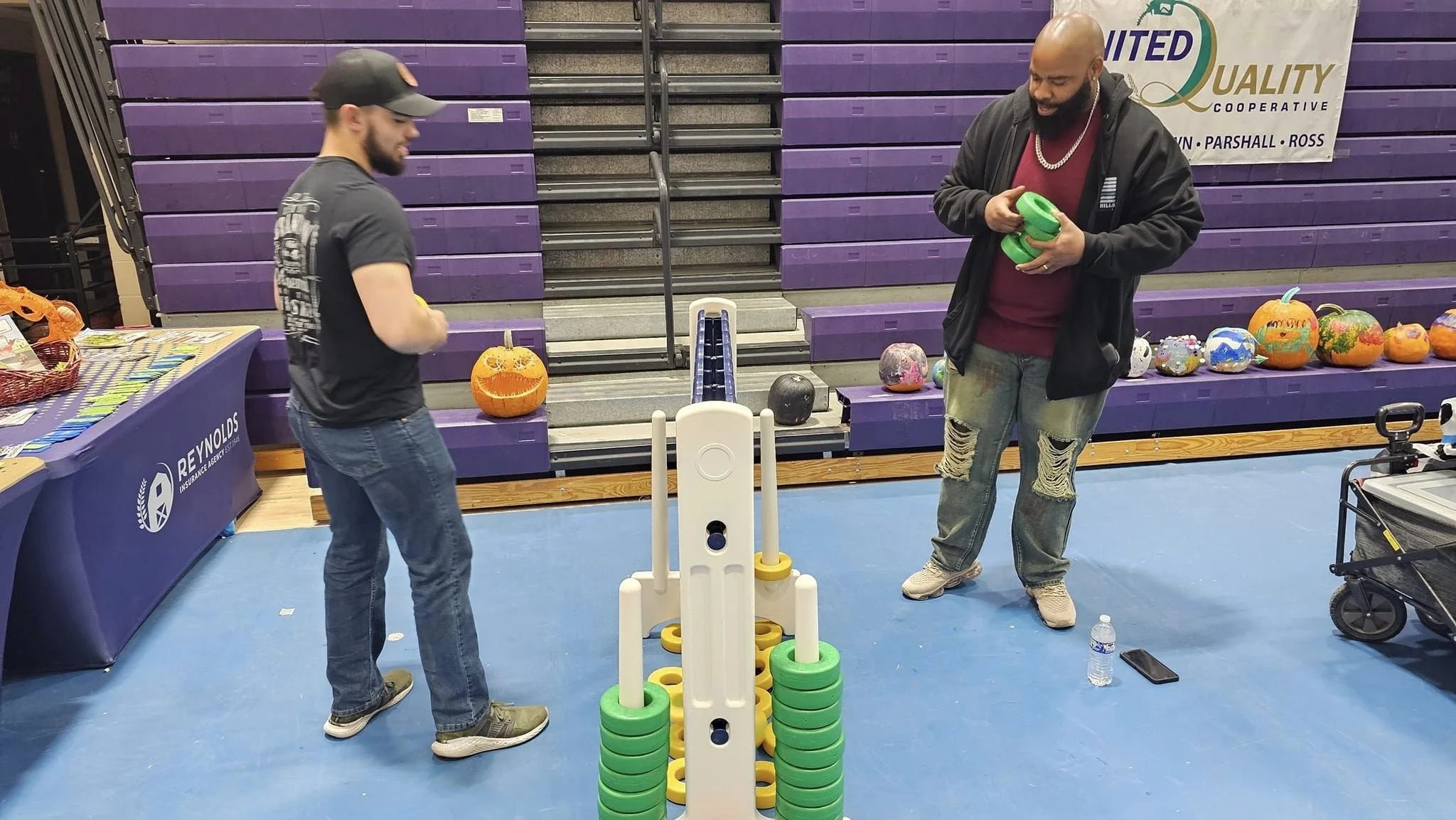 Two men playing a game of giant Jenga in a gymnasium gym, with purple bleachers and Halloween decorated pumpkins behind them.
