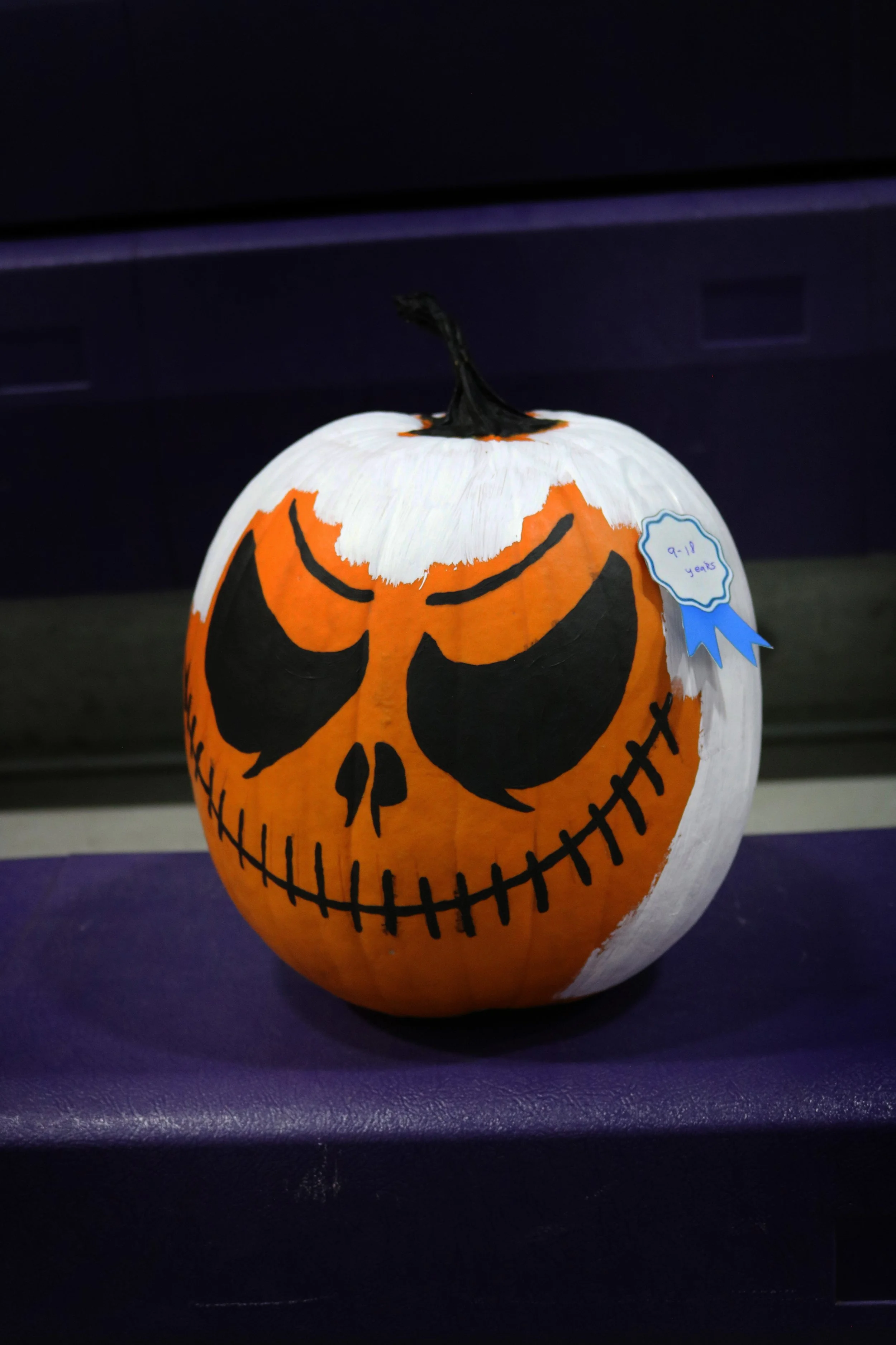 A white pumpkin decorated with a painted orange, black, and white Halloween face, resembling a spooky Jack-o'-lantern with a black stem and a blue ribbon sticker indicating it is for ages 9-12.