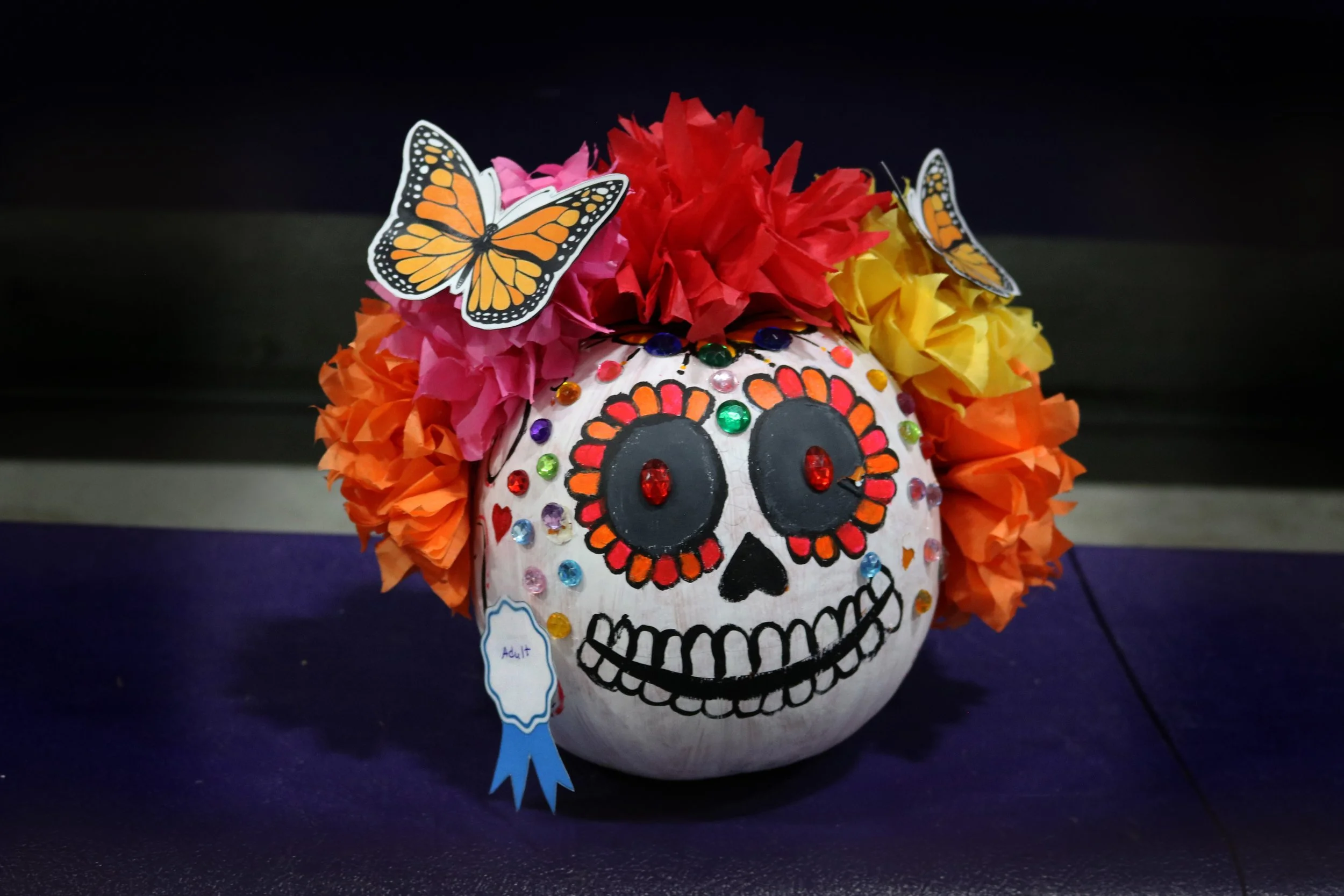Decorative skull painted in traditional Mexican calavera style, decorated with colorful flowers, stickers, and butterflies, positioned on a purple surface.