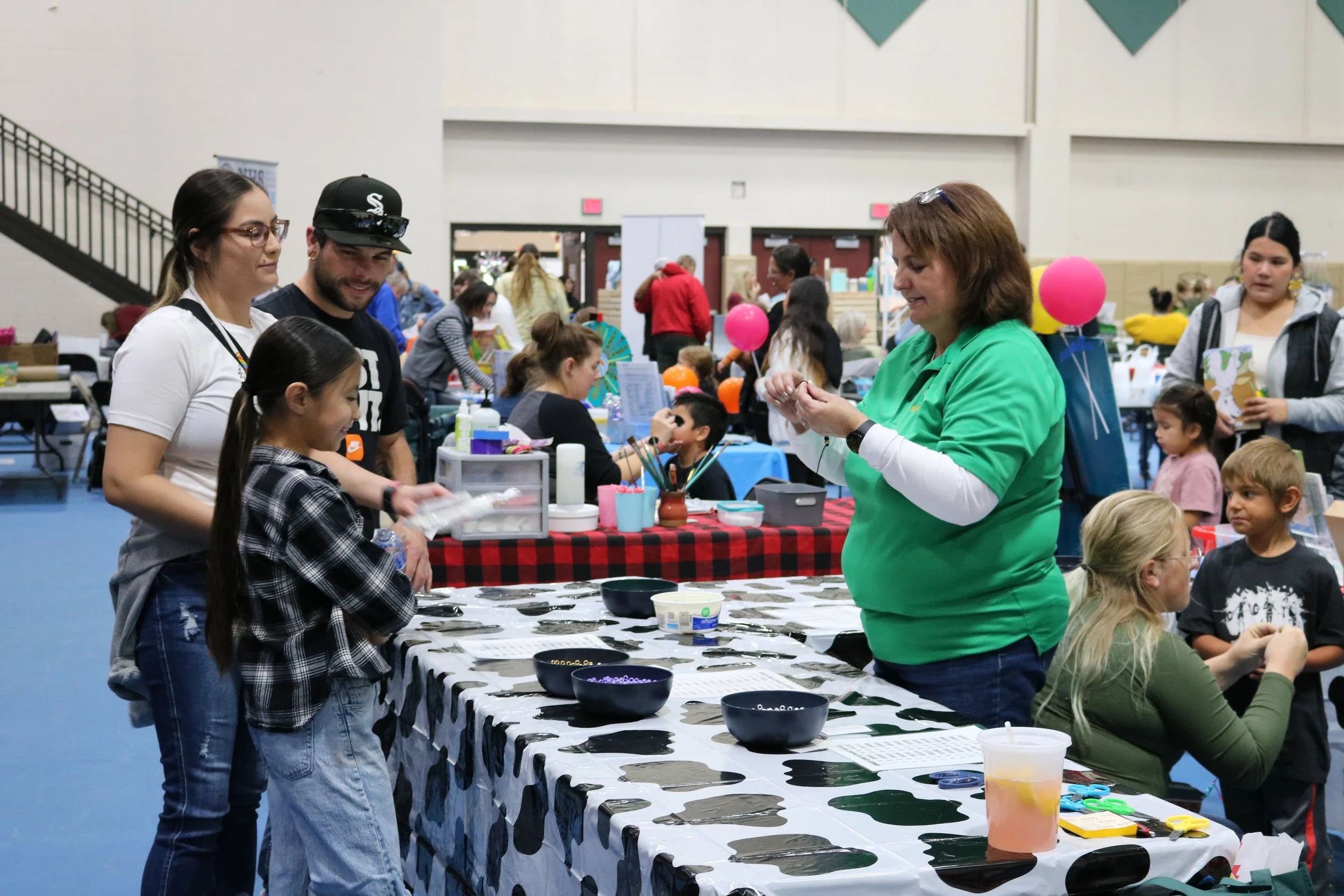 A group of people at an indoor craft fair, with a woman in a green shirt demonstrating something at a table covered with black and white patterns, and several children and adults engaging in activities around.