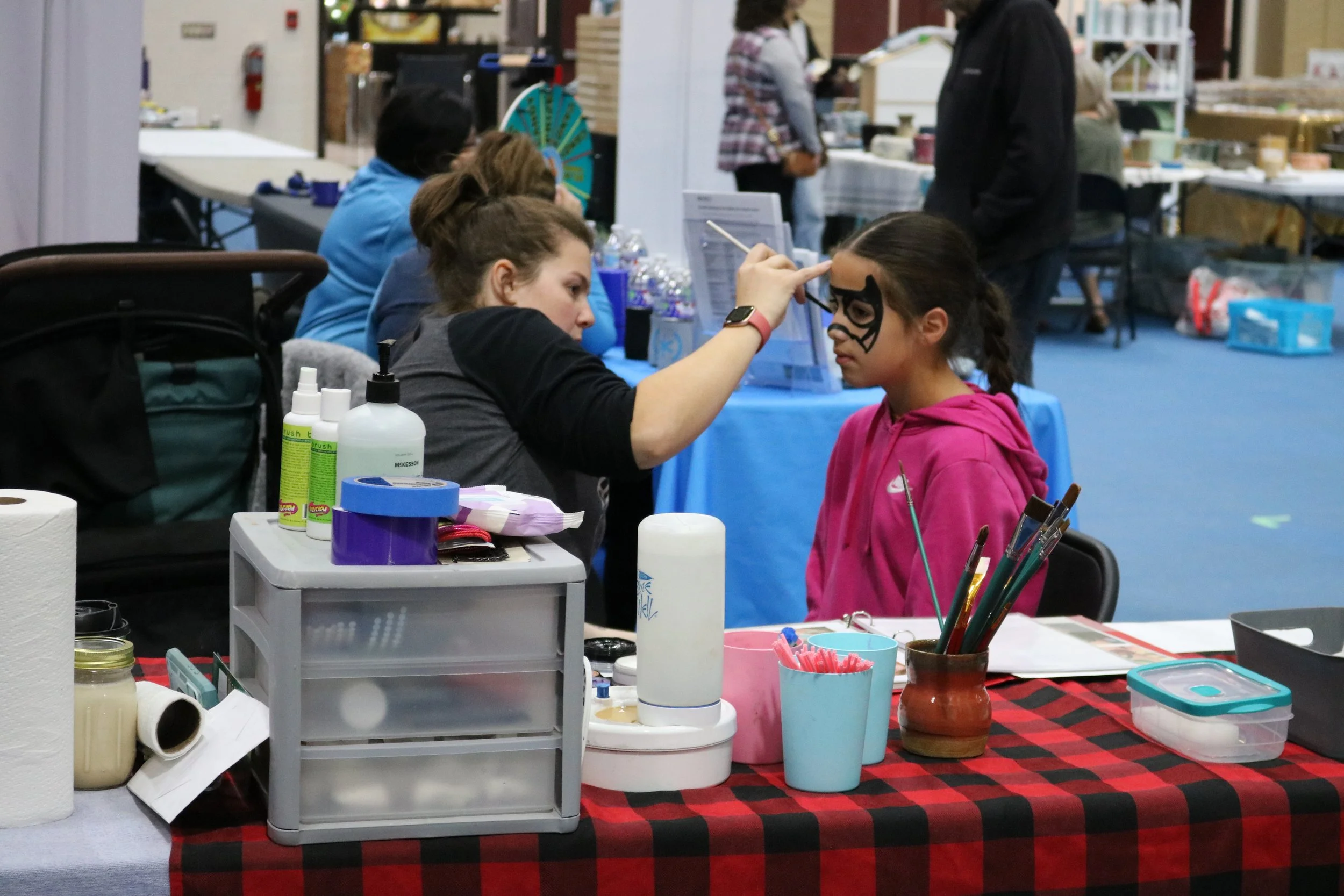 A girl in a pink hoodie having her face painted by a woman at a booth with face painting supplies, including brushes, paint, and water bottles, with other booths and people in the background.