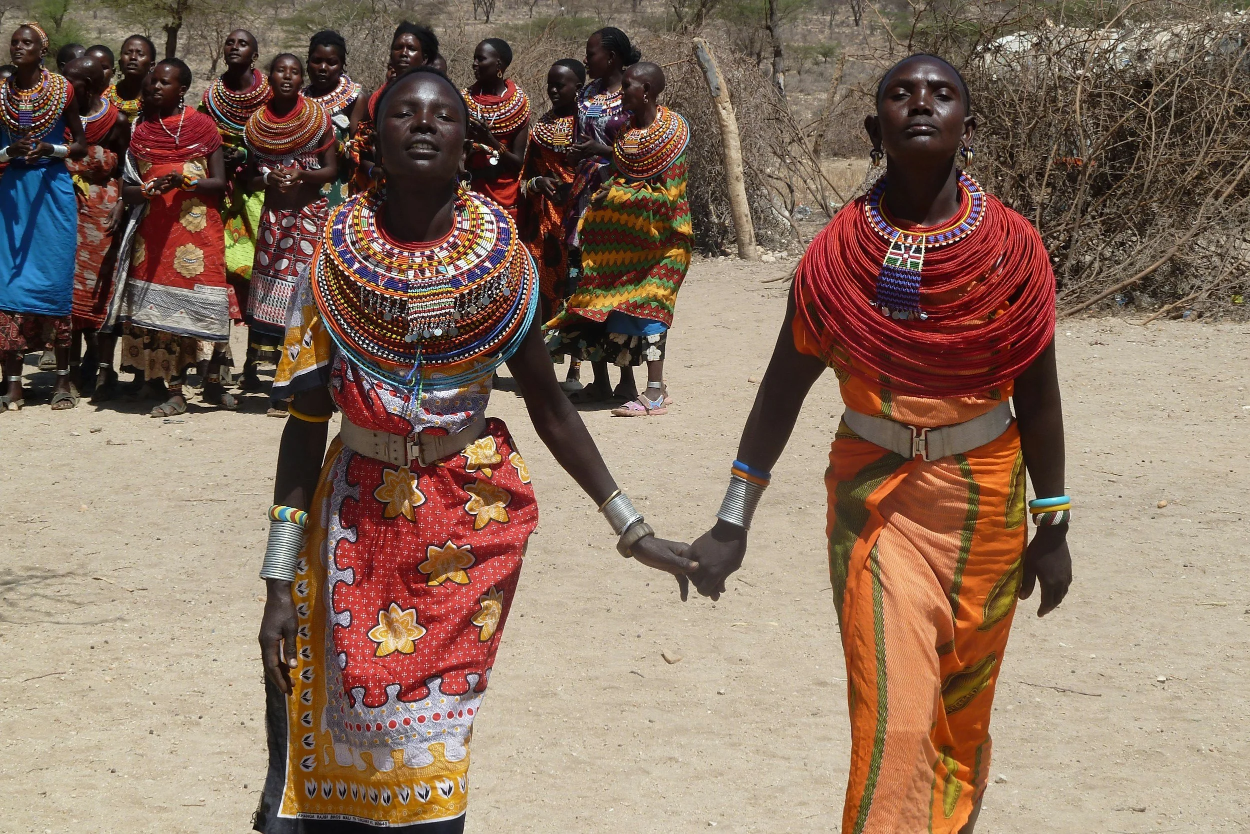 Two women dressed in traditional Maasai clothing holding hands in the foreground, with a group of women in colorful attire behind them, in a dry, outdoor setting.