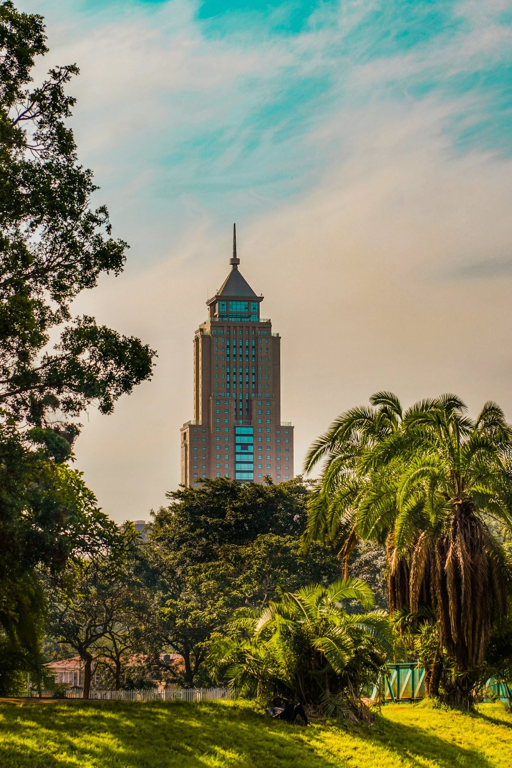 A tall skyscraper with a pointed top seen through trees, with a park featuring lush green grass and palm trees in the foreground.