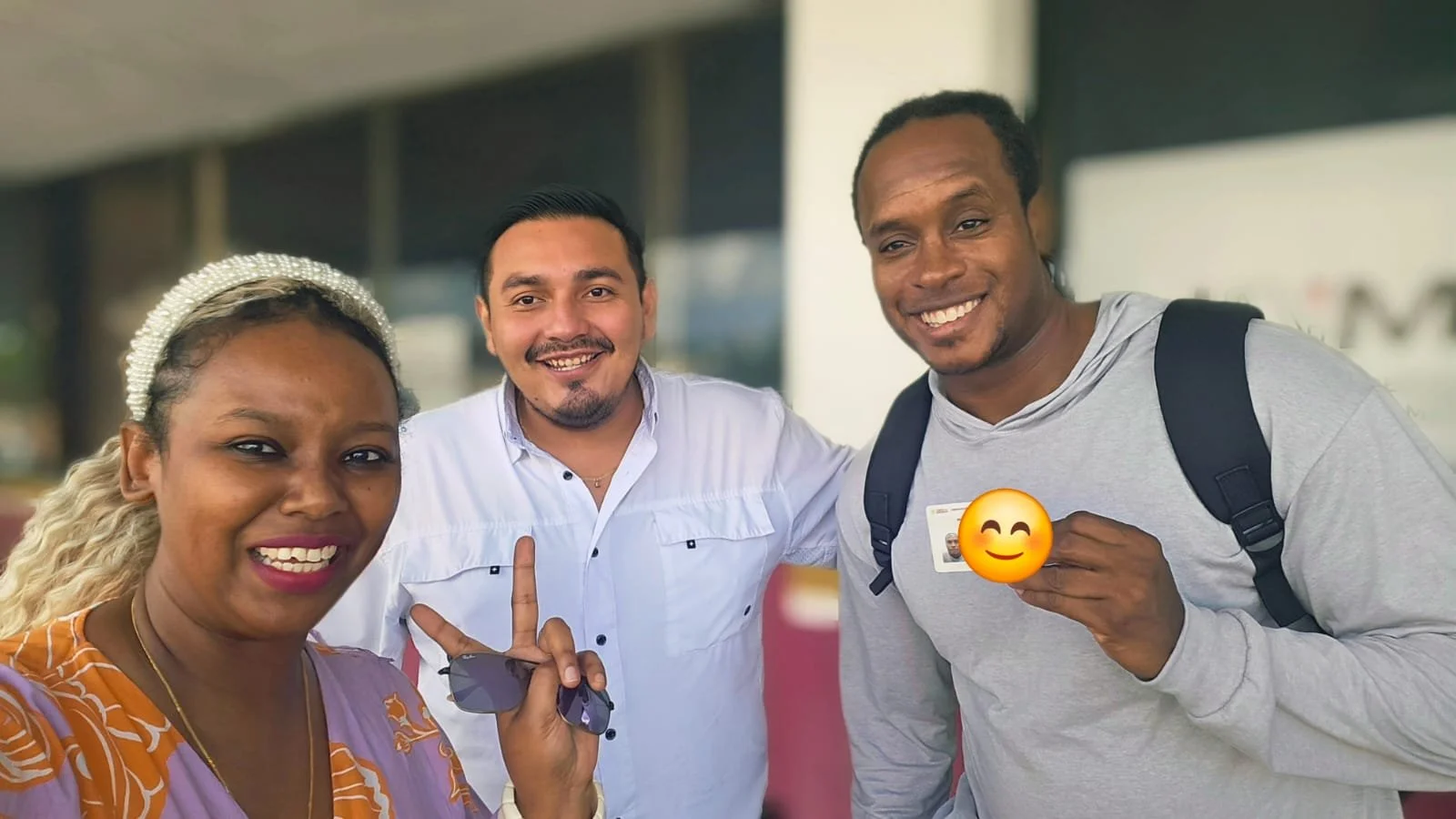 Three smiling people taking a selfie, with one holding an ID card, outdoors near a building.