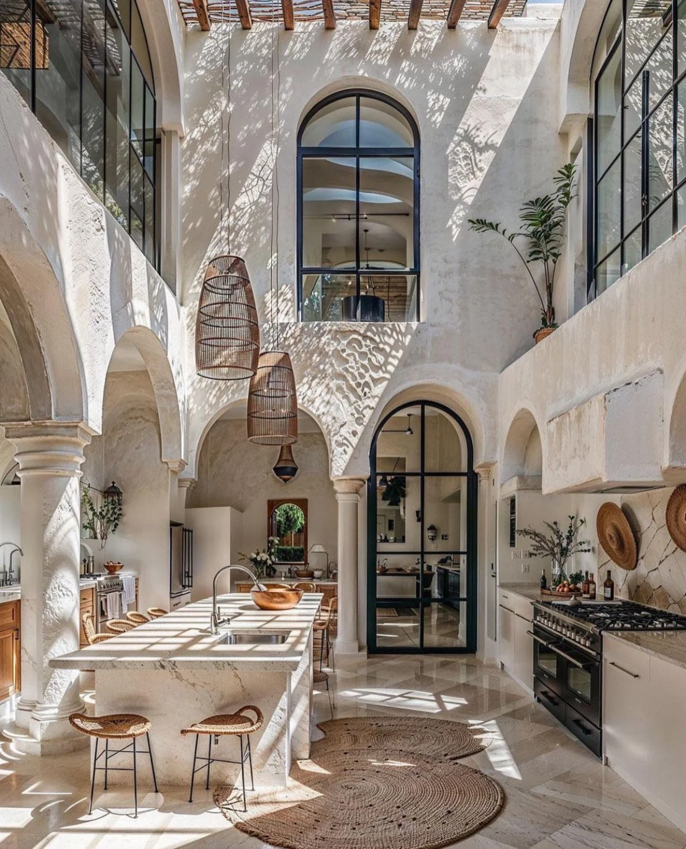 Interior view of a bright, airy kitchen with high ceilings, large arched windows, and floating wooden pendant lights. white cabinetry, a marble kitchen island, and natural woven rugs are visible, with sunlight casting shadows through a pergola above.