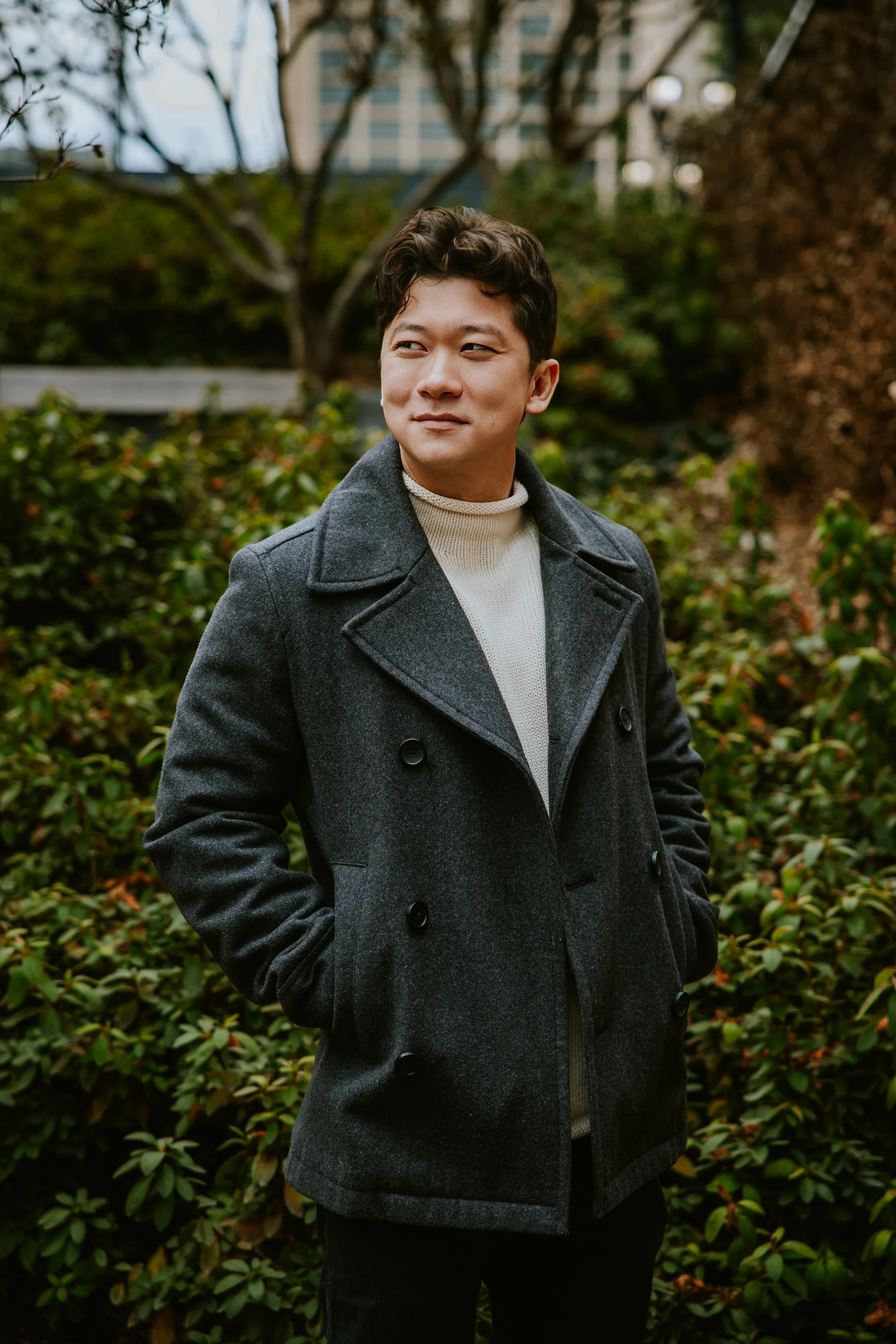 A man standing outdoors in a park, wearing a gray coat and a white sweater, with hands in coat pockets, looking to his left.