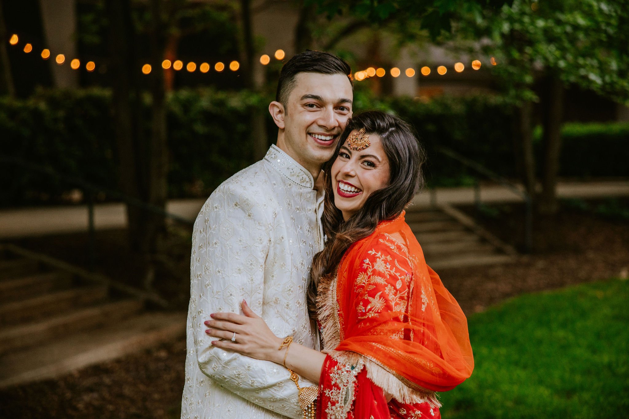 A smiling couple dressed in traditional Indian wedding attire, standing outdoors with string lights and trees in the background.