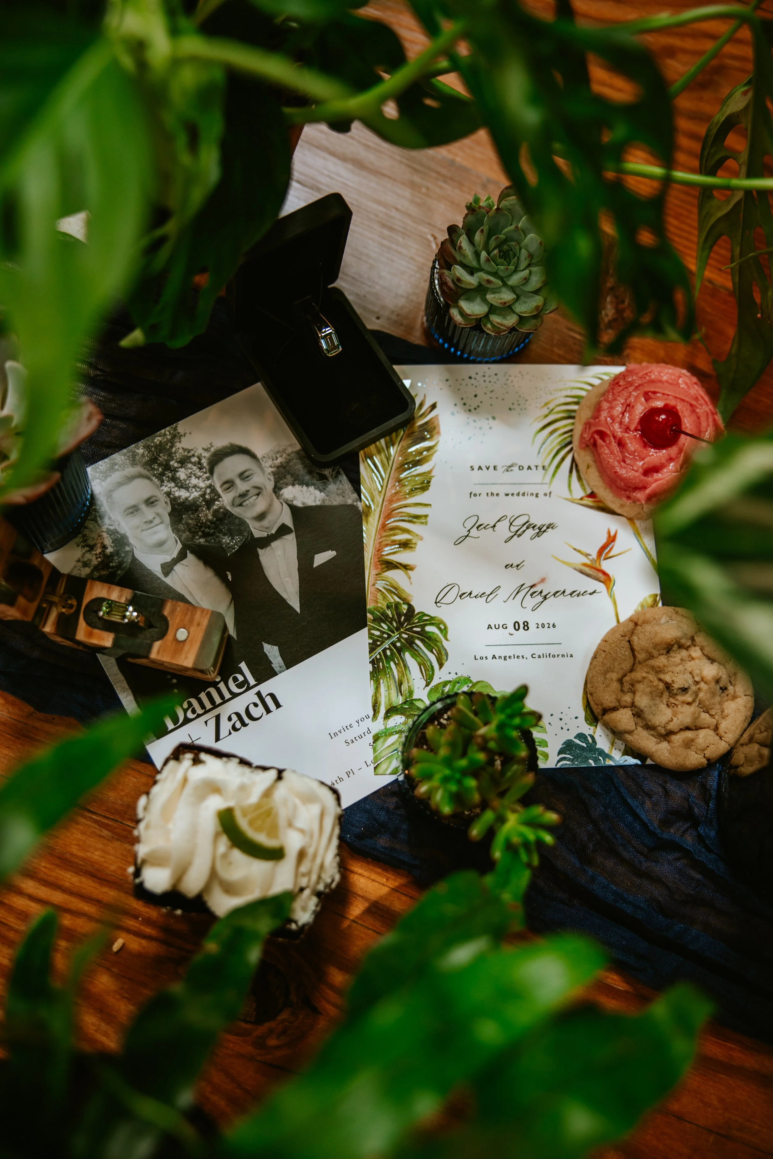Horizontal photo of wedding invitation flatlay featuring wedding rings and Crumbl cookies