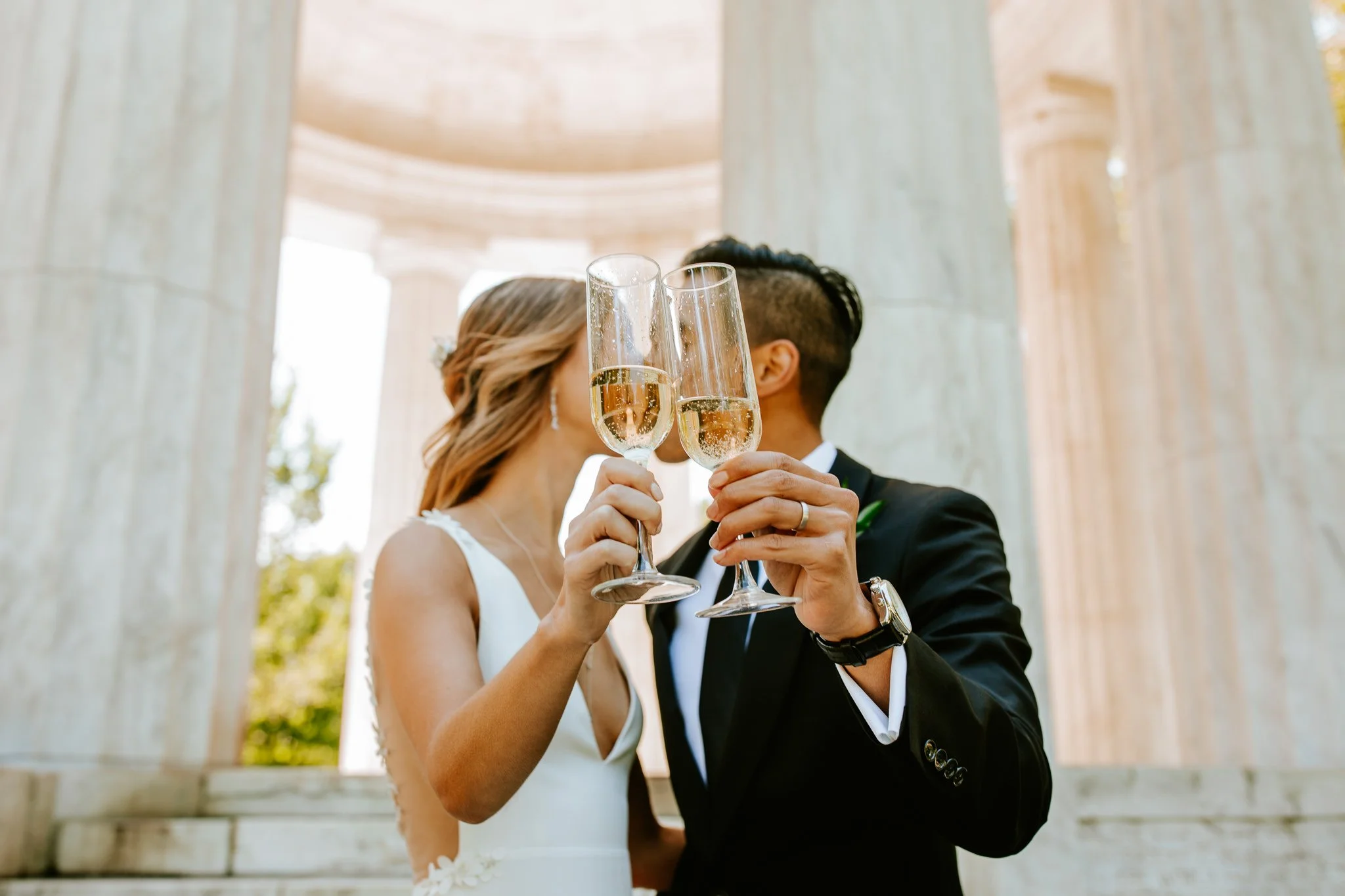 A bride and groom at their wedding ceremony raising champagne flutes for a toast, standing closely together in front of classical columns.