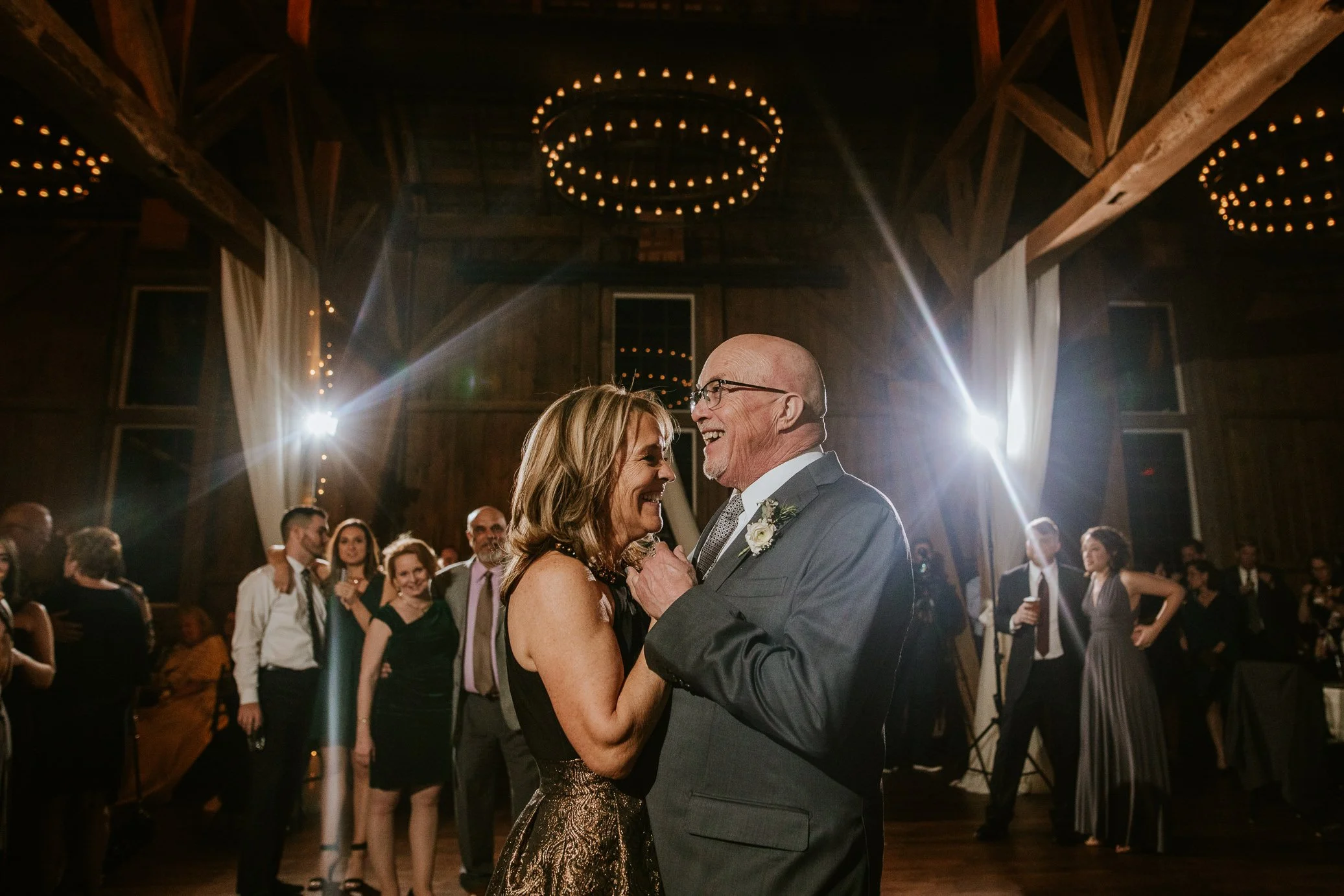 A couple shares a dance at a wedding reception, surrounded by guests in a rustic barn with warm lighting and circular chandeliers.
