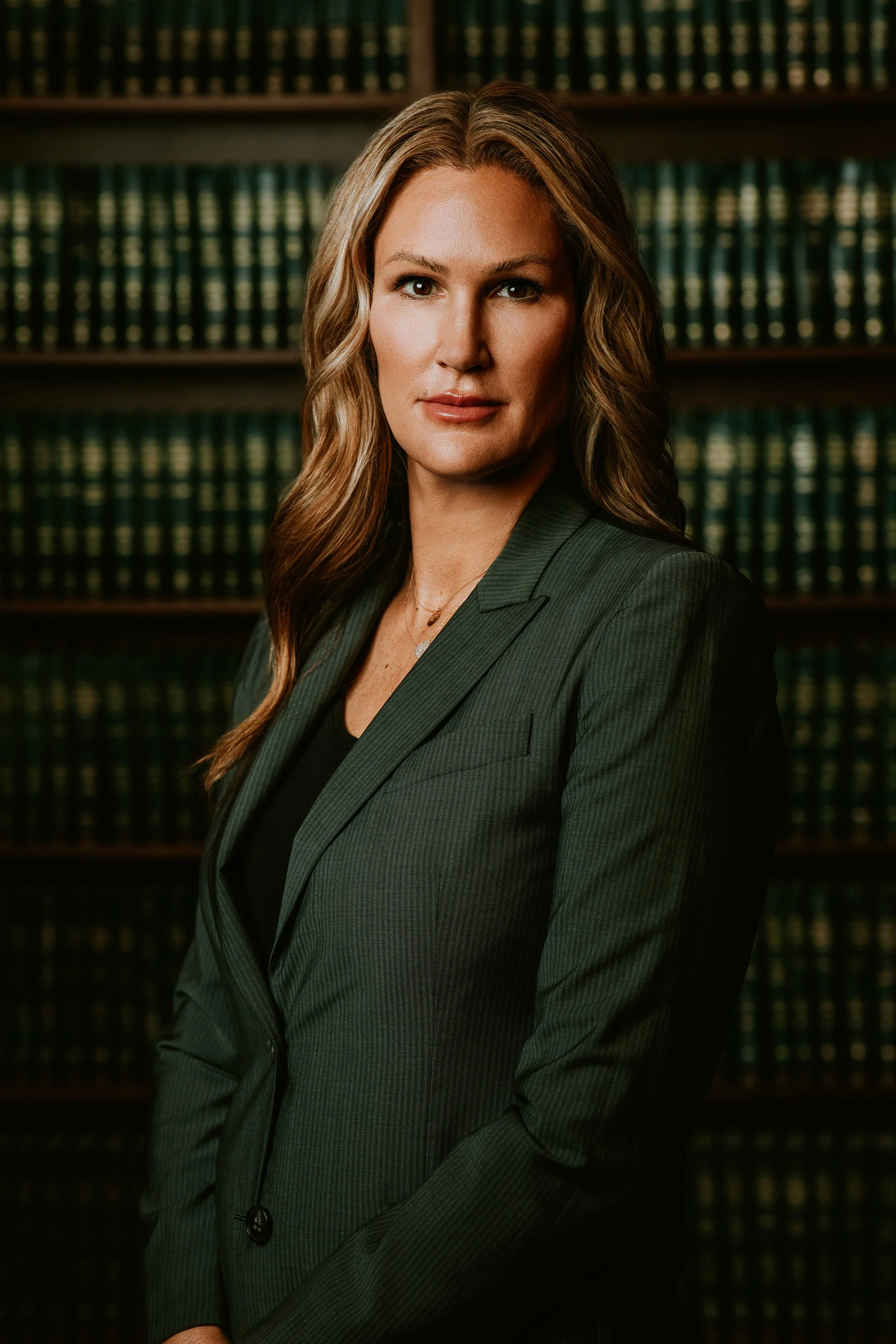A woman with long wavy brown hair wearing a dark green pinstripe suit standing in front of shelves filled with law books.