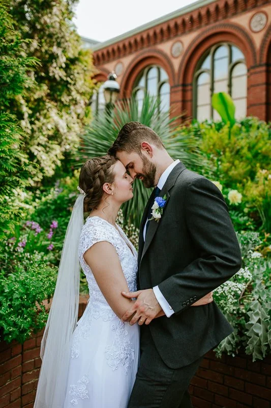 A newlywed couple sharing a tender moment with foreheads touching, surrounded by lush greenery and flowers in an outdoor garden setting.