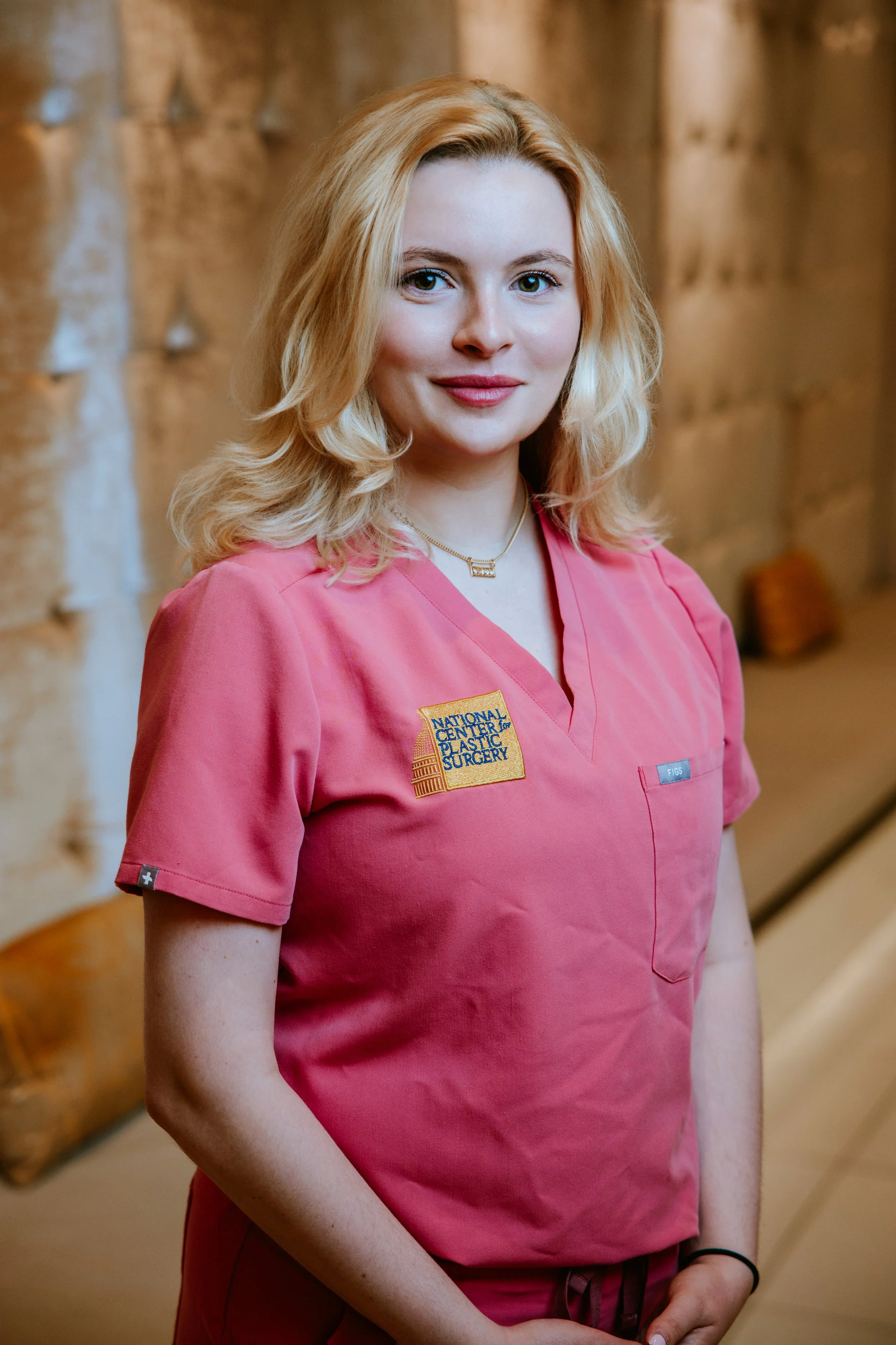 A young woman with blonde hair wearing pink medical scrubs with a patch that says 'National Center for Plastic Surgery' on the chest, standing indoors against a blurred background with exposed brick wall.