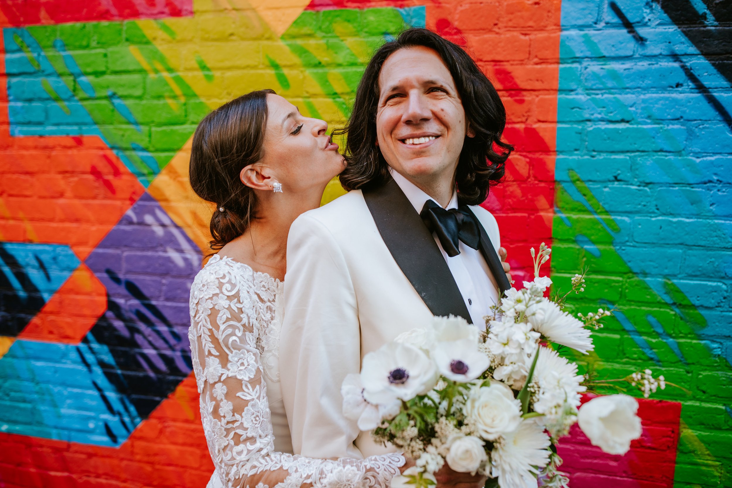 A woman in a wedding dress and a man in a tuxedo pose for a photo in front of a colorful mural wall. The woman is kissing the man's cheek and holding a bouquet of flowers.