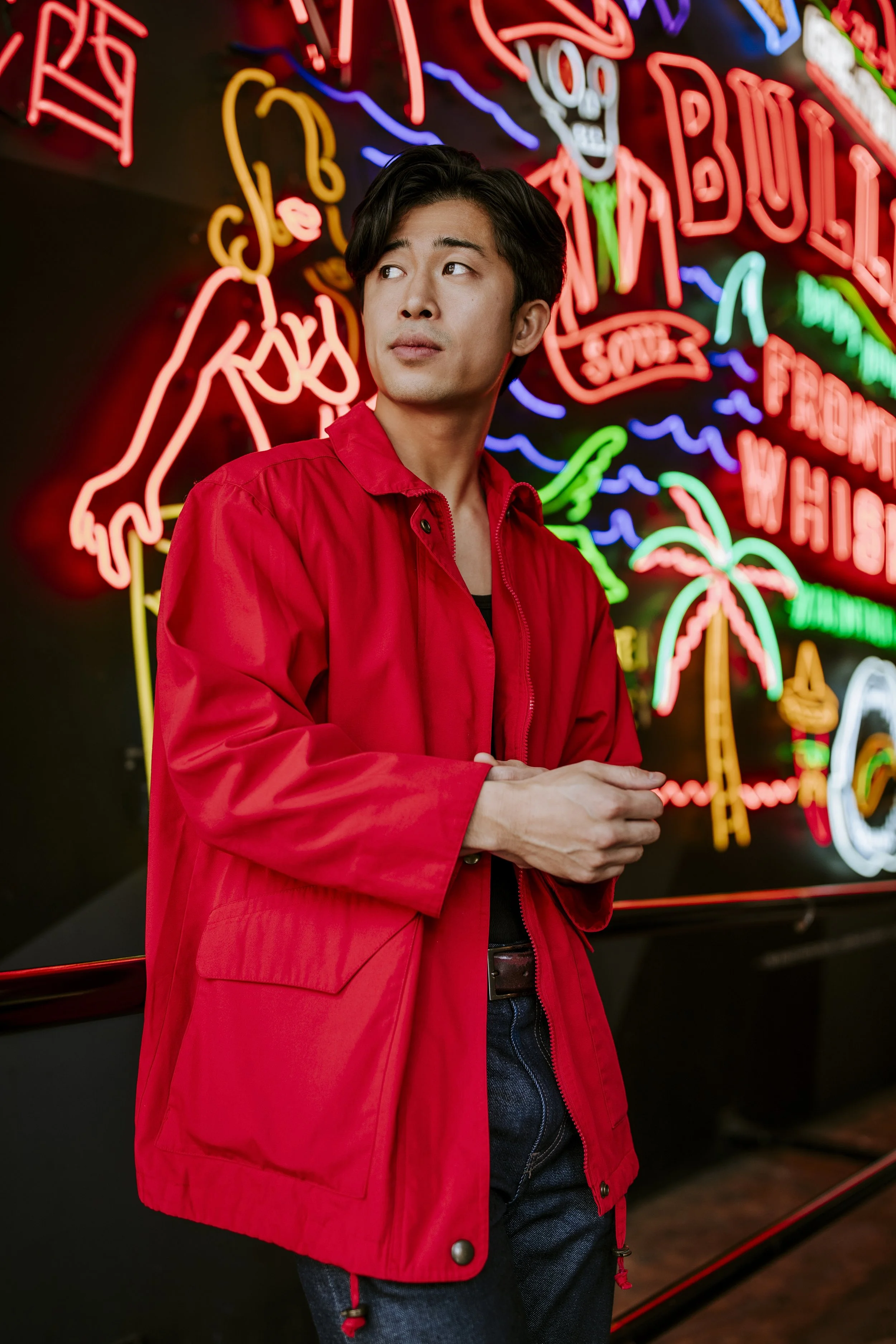 A young man in a red jacket standing in front of neon signs with various colorful graphics and text at night.
