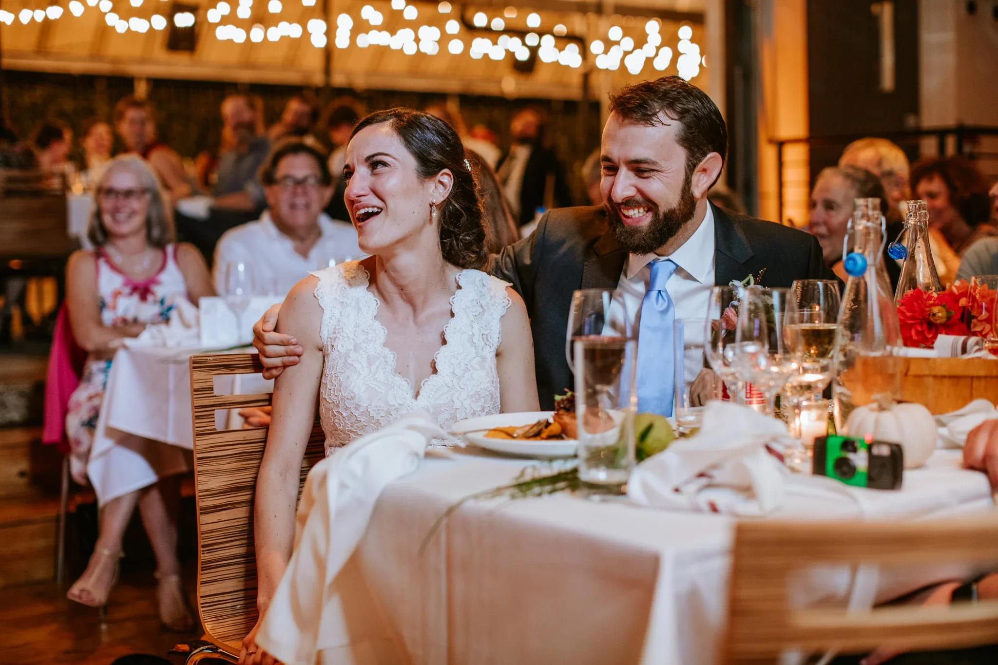A bride and groom sitting at a table during a wedding reception, smiling and laughing, with guests in the background. The bride is wearing a white lace dress, and the groom is in a dark suit with a light blue tie.