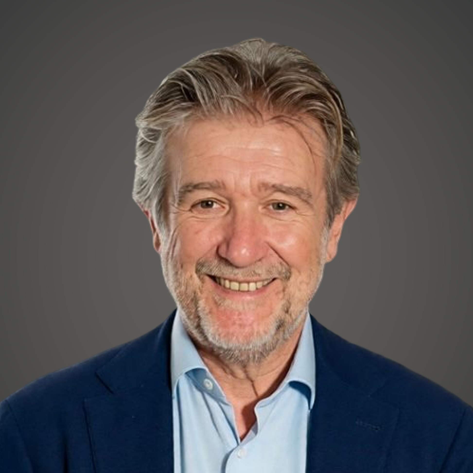 Close-up of a smiling middle-aged man with gray hair and a beard, wearing a suit jacket and dress shirt, outdoors with greenery and a white support structure in the background.