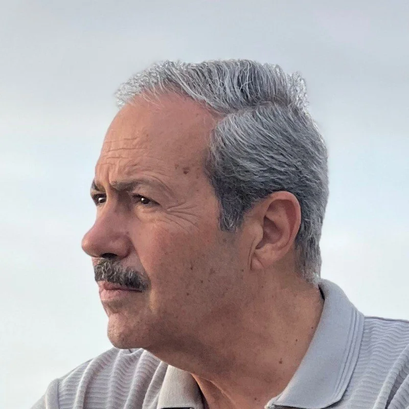 Side view of an older man with gray hair and a mustache, looking serious outside with a cloudy sky background.